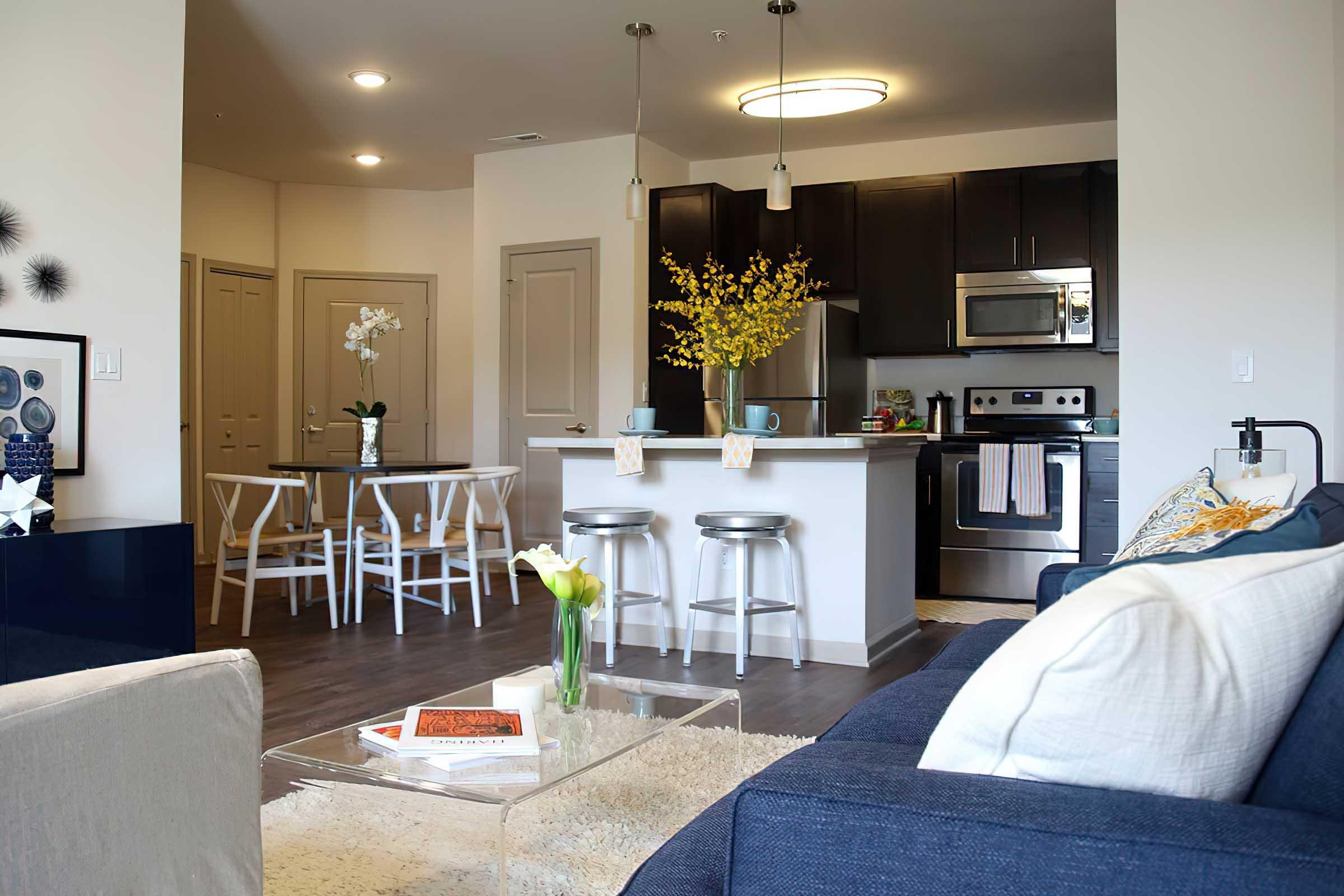 Modern apartment interior featuring a living area with a blue sofa and a glass coffee table. In the background, a kitchen with dark cabinets and stainless steel appliances is visible. A dining table with white chairs is set, and decorative elements like flowers are placed throughout the space, giving a cozy atmosphere.