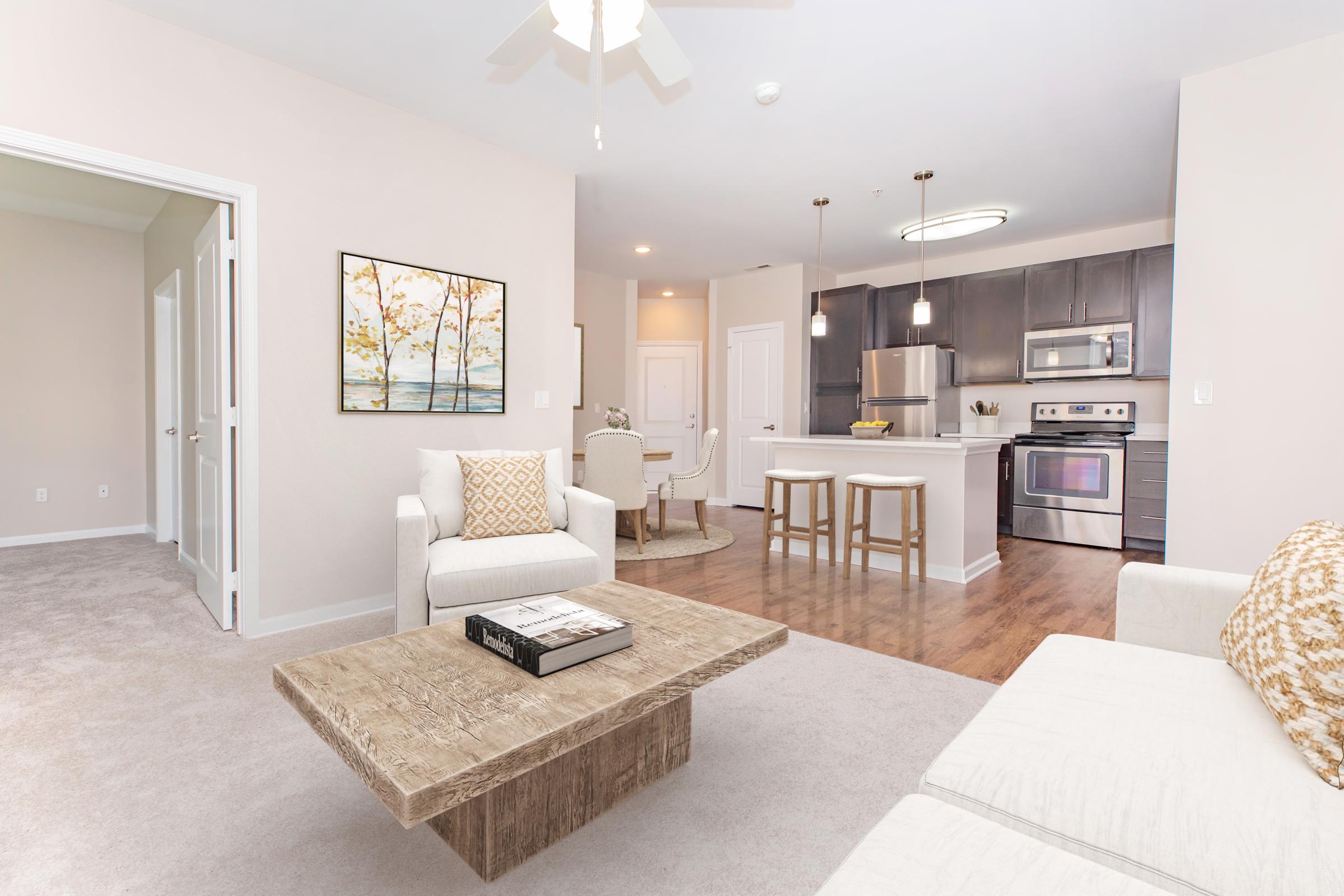A modern living room featuring a light-colored sofa and a natural wood coffee table. The space has neutral walls and carpet, with an art piece on the wall. An adjacent kitchen showcases dark cabinetry and stainless steel appliances, creating a cozy and inviting atmosphere.