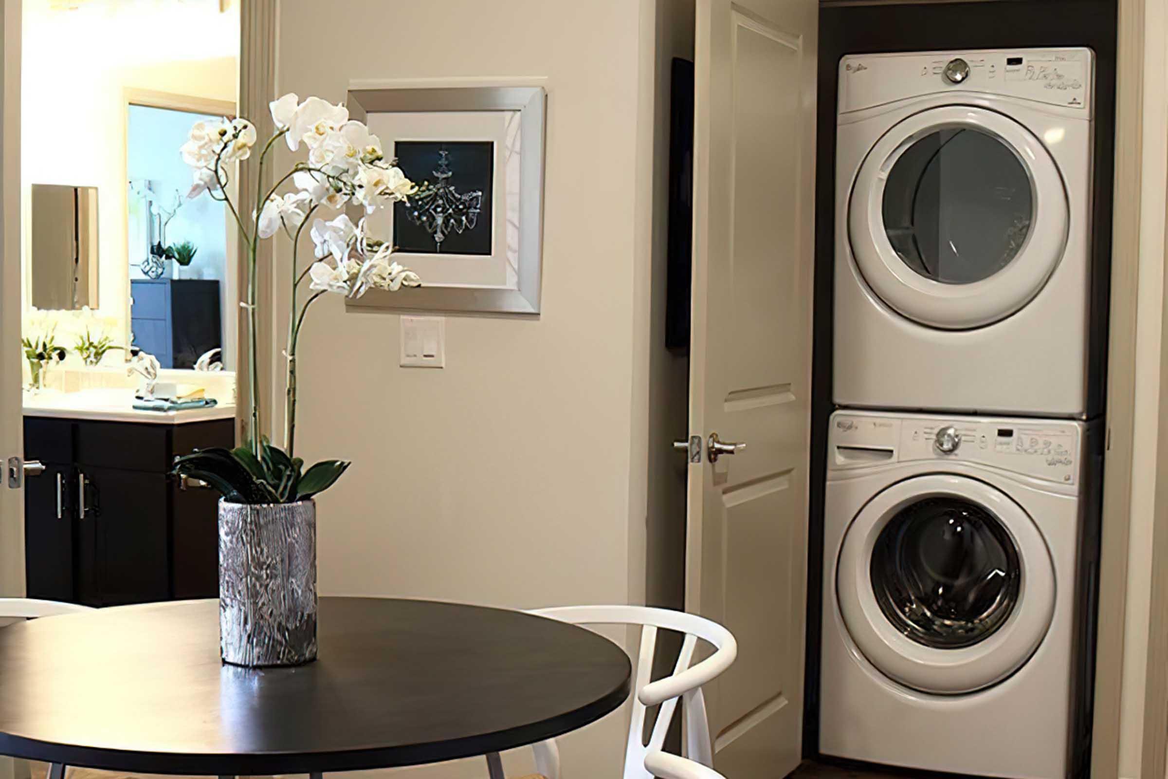 A modern laundry area featuring stacked washing machines beside a doorway. A round dining table with white chairs is in the foreground, adorned with a vase of white orchids. In the background, a bathroom is visible with soft lighting and elegant decor.