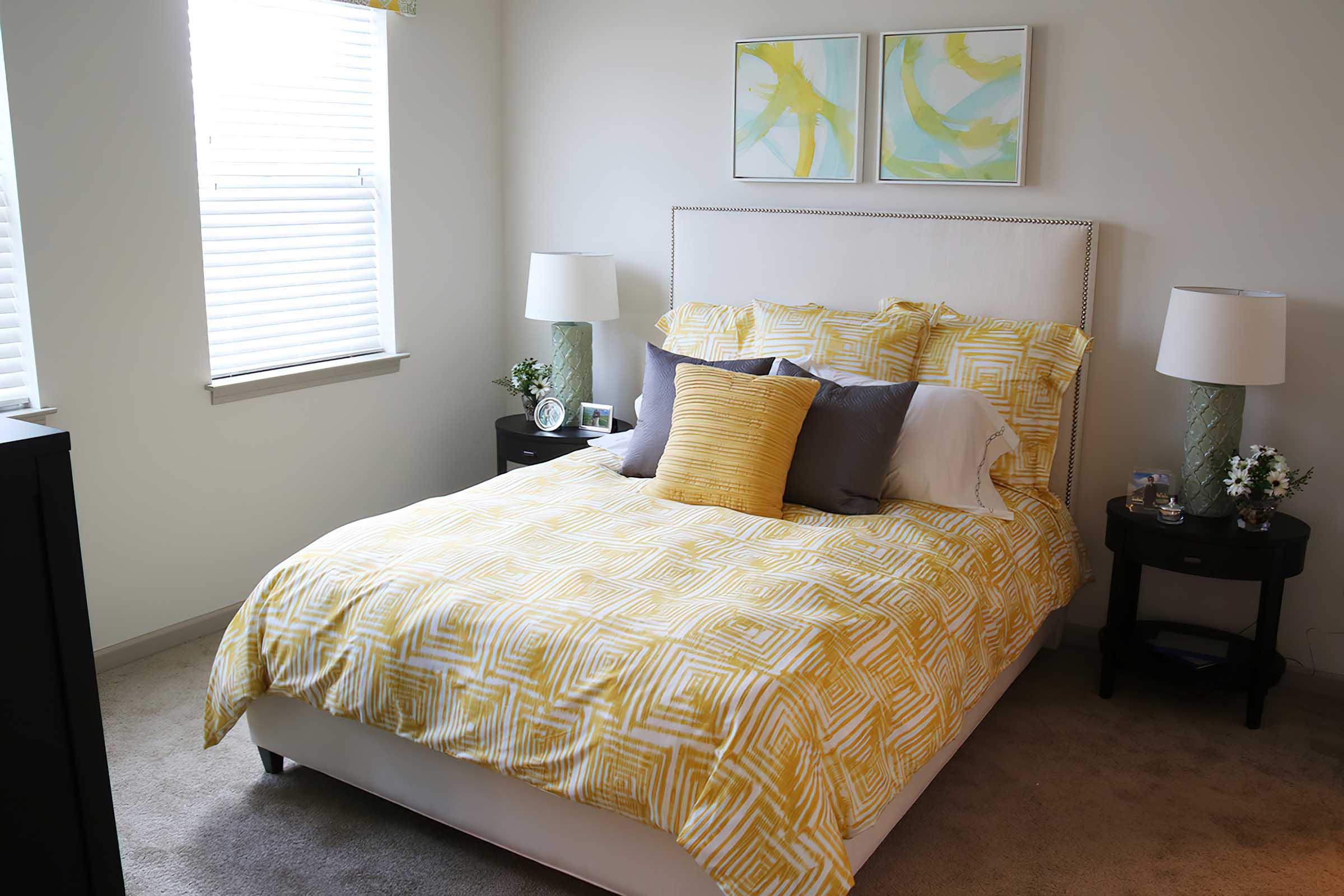 A cozy bedroom featuring a white bed with a yellow and white patterned duvet cover. The bed is adorned with gray and yellow pillows and flanked by two nightstands with lamps. There are two abstract paintings on the wall and a window with sheer curtains allowing natural light to brighten the space.