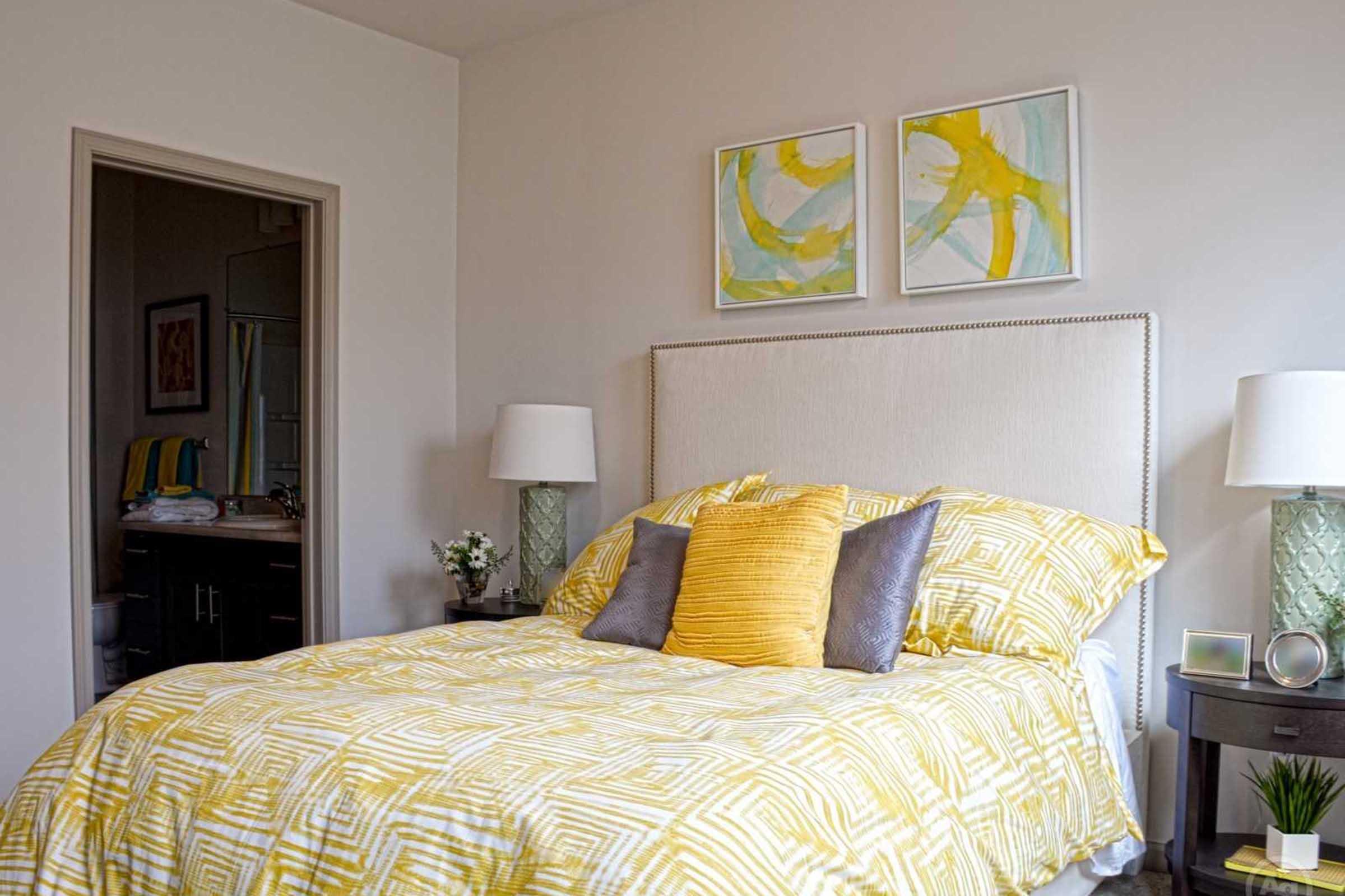 A cozy bedroom featuring a yellow and white patterned bedspread with multiple accent pillows. Two bedside lamps are on either side of the bed, and there are abstract paintings above the headboard. A bathroom is visible in the background through an open door.