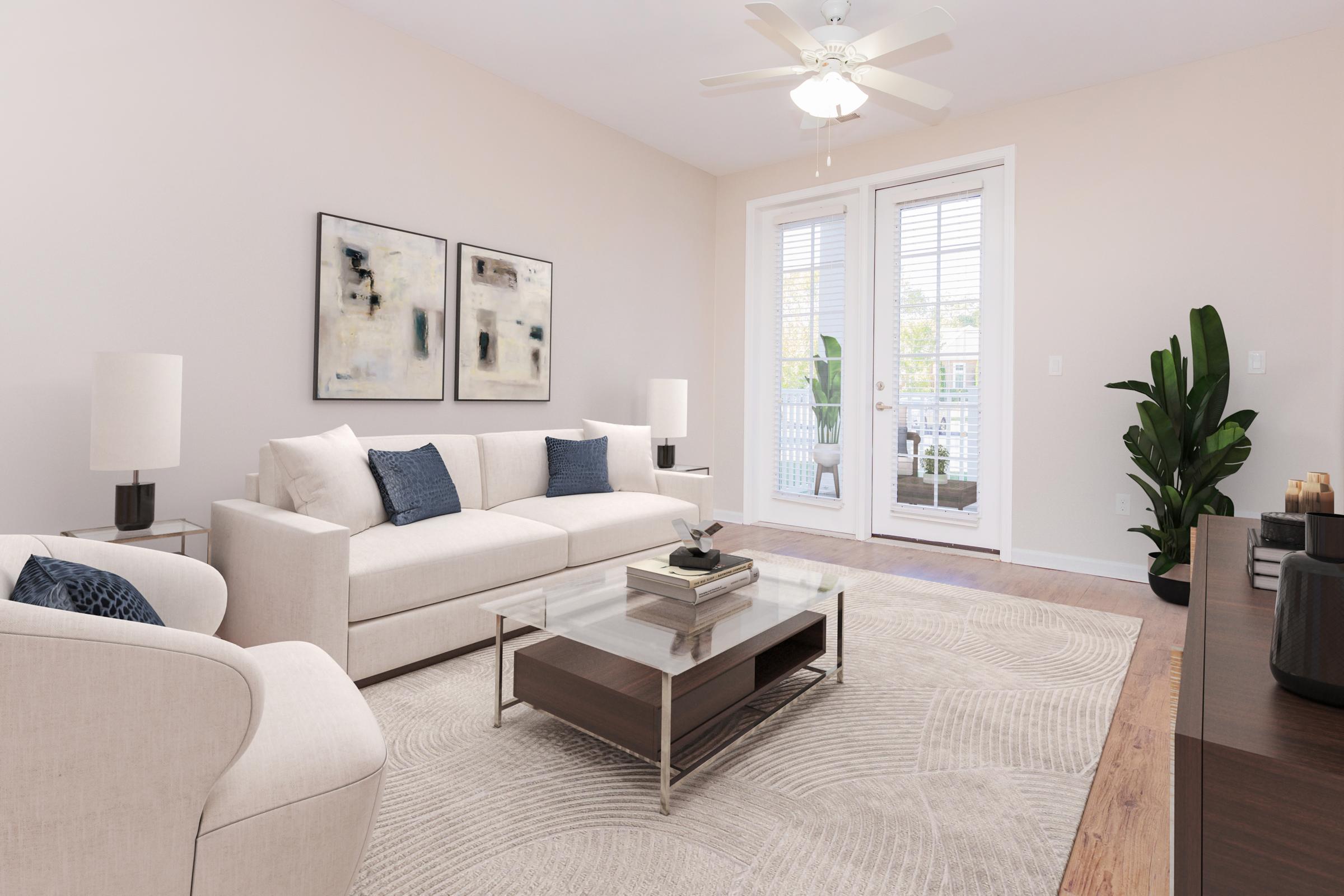 A modern living room featuring a light-colored sofa with blue accent pillows, a glass coffee table, and two white lamps. There are abstract artworks on the wall, a ceiling fan above, and large doors leading to a balcony. A green plant adds a touch of nature to the space, which has warm wood flooring.