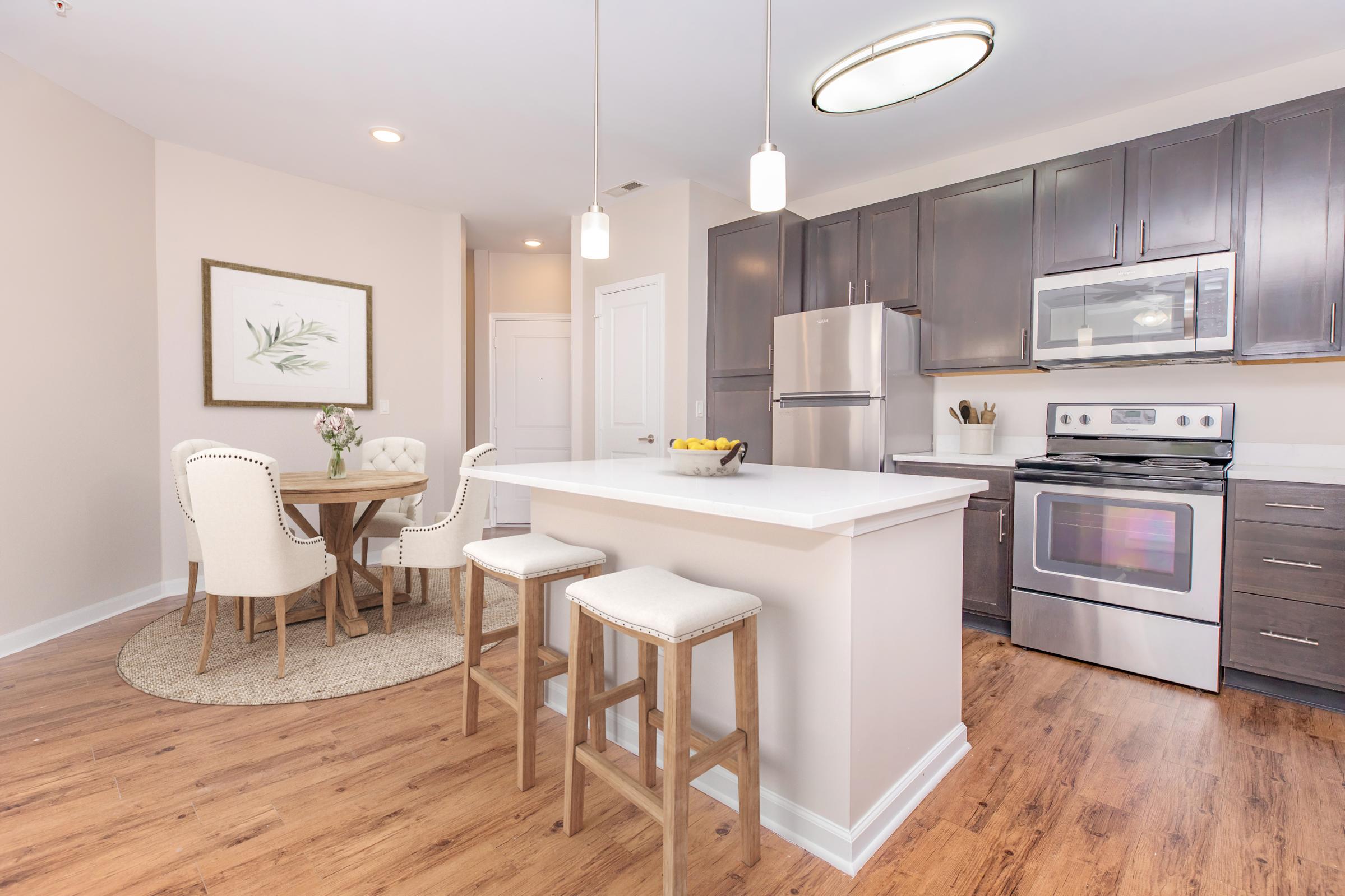 Modern kitchen with dark wood cabinets, stainless steel appliances, and a white countertop. There is a small dining area with a round table and four upholstered chairs. Pendant lights hang above the counter, and a tasteful framed artwork is displayed on the wall. A decorative rug is under the table on a warm wood floor.