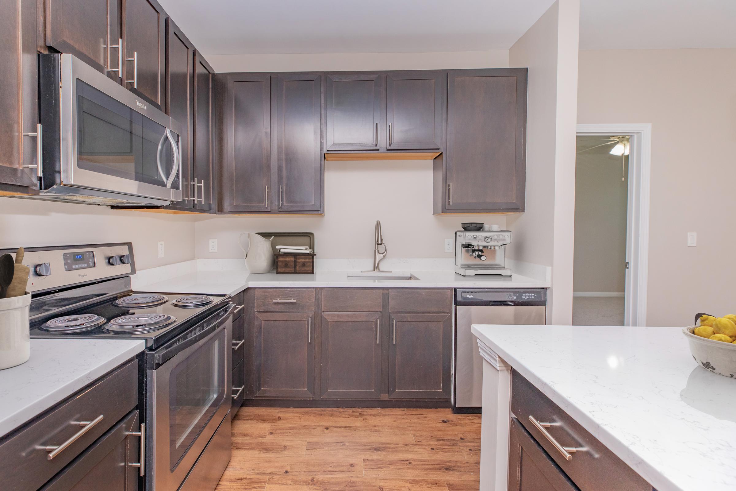 Modern kitchen featuring dark cabinetry, stainless steel appliances including an oven and microwave, a white countertop, and hardwood flooring. A sink is placed against the wall with decorative items nearby. Bright, neutral walls enhance the space's overall aesthetic.