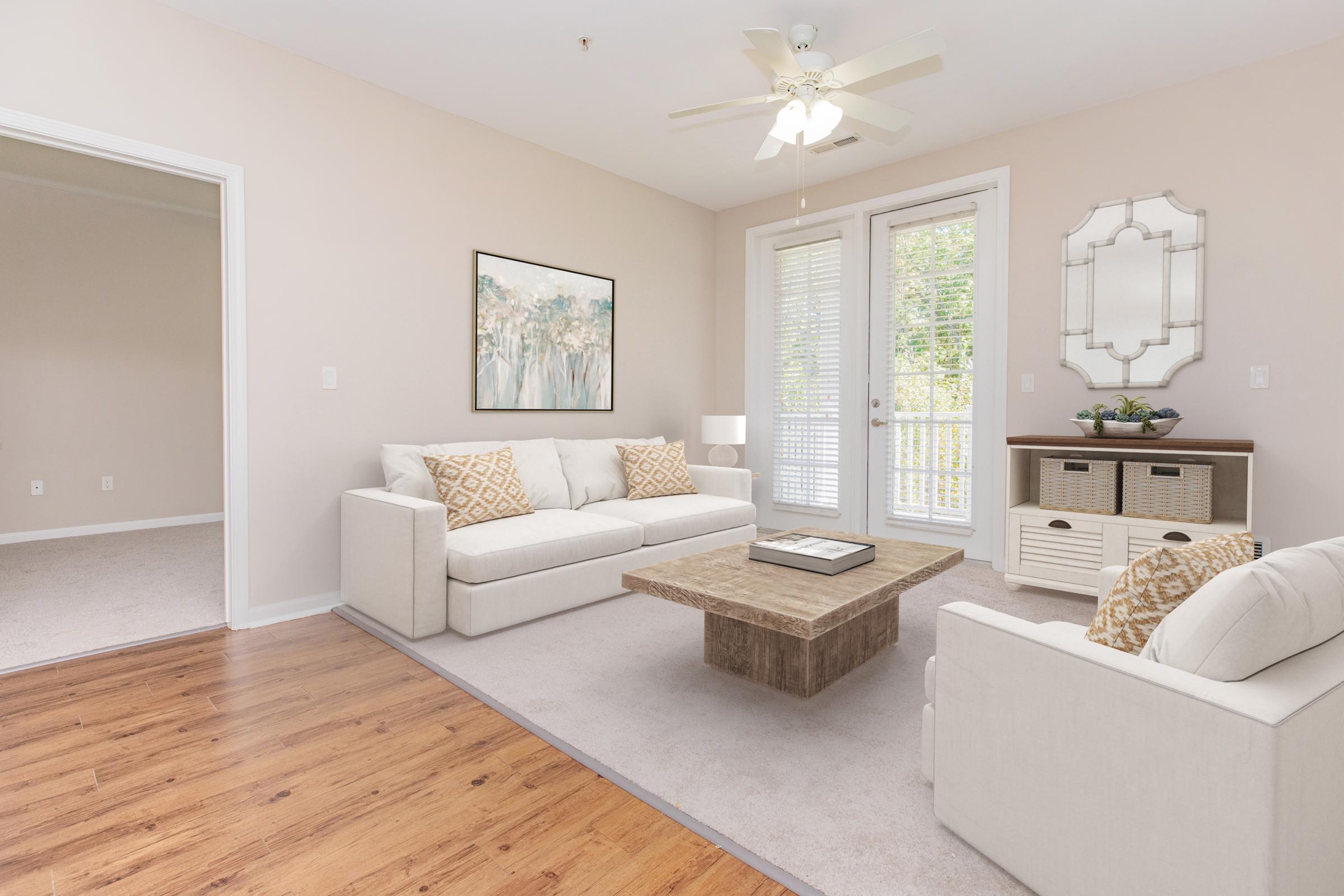 A bright living room featuring a light-colored sofa with patterned cushions, a coffee table in the center, and large windows with sliding doors that bring in natural light. There is a mirror and decorative items on a console table against the wall. The flooring is wooden, adding warmth to the space.