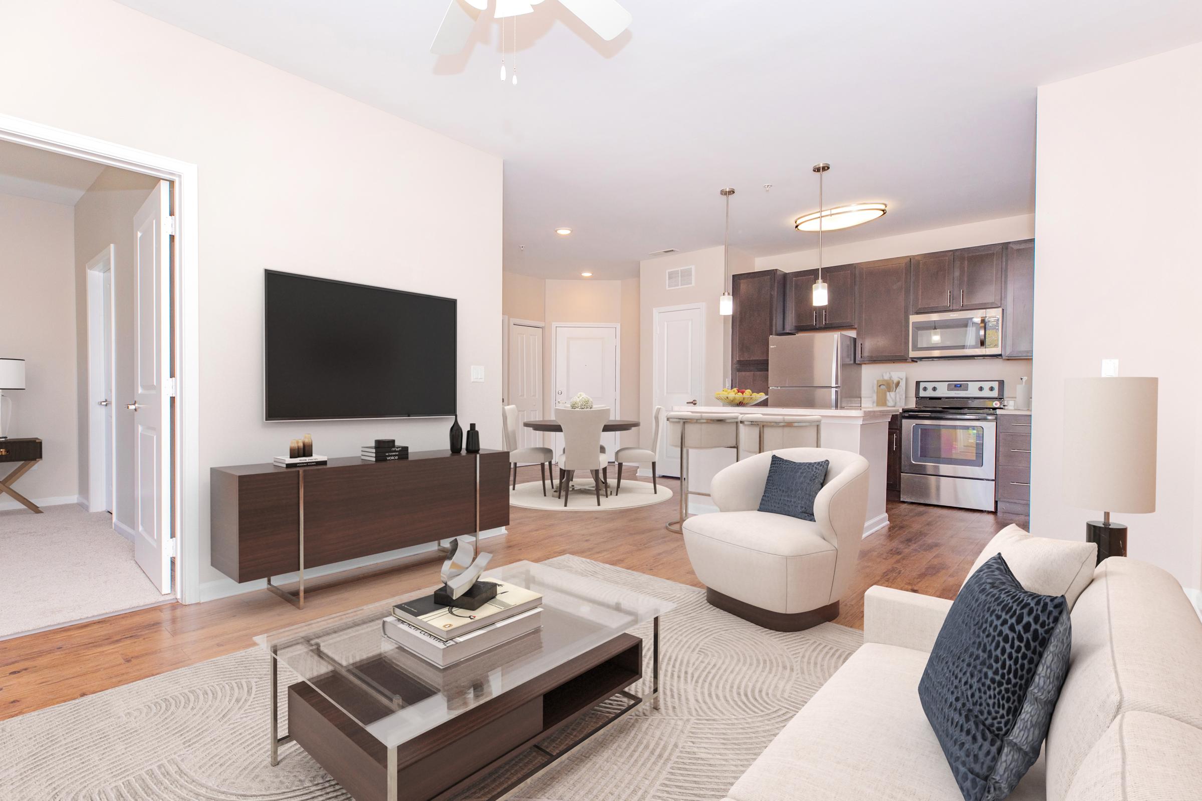 A modern living room featuring a light-colored sofa with blue cushions, a glass coffee table, and a television mounted on the wall. The room has wooden flooring and a ceiling fan, with a dining area visible in the background that includes a round table and chairs. Soft, neutral colors create a welcoming atmosphere.