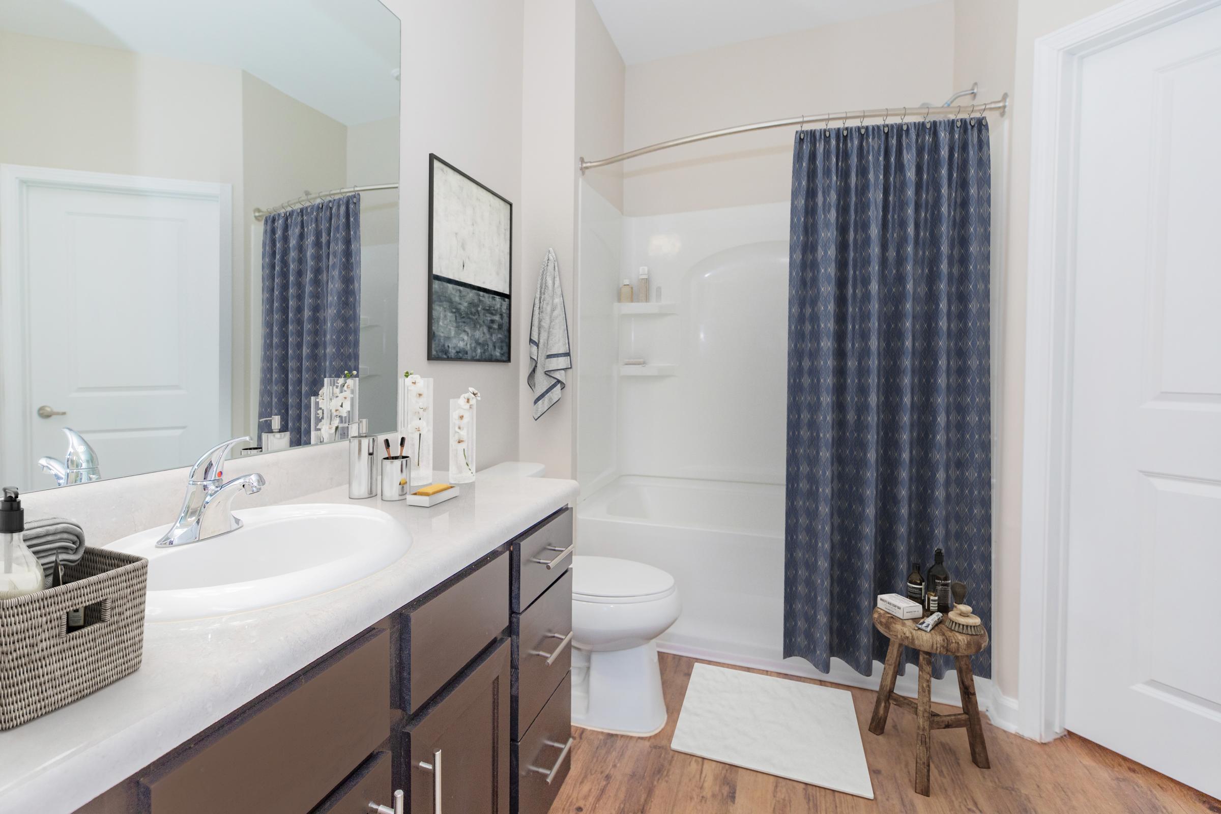 A modern bathroom featuring a white sink with a chrome faucet, a large mirror, and dark brown cabinetry. There's a shower with a navy blue curtain, a white bathtub, and a small decorative stool. The floor is wooden, and accessories such as toiletries are neatly arranged on the countertop. The walls are painted in a light color, creating a bright, inviting atmosphere.