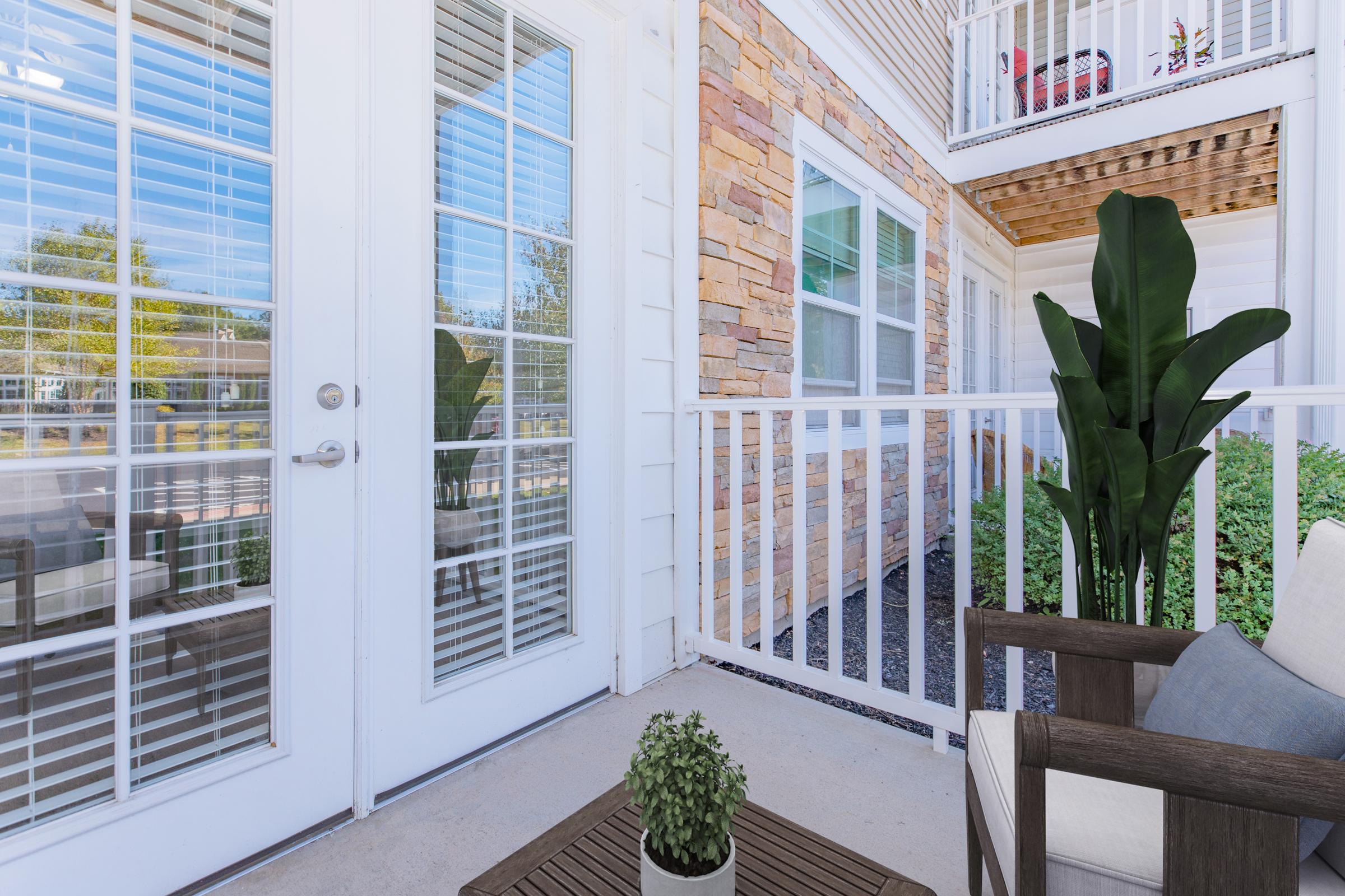 A cozy outdoor space featuring a small table with a potted plant, a comfortable chair, and large glass doors with blinds. The backdrop includes a textured stone wall and a balcony above, with a clear blue sky visible through the glass. The area is well-lit and inviting.