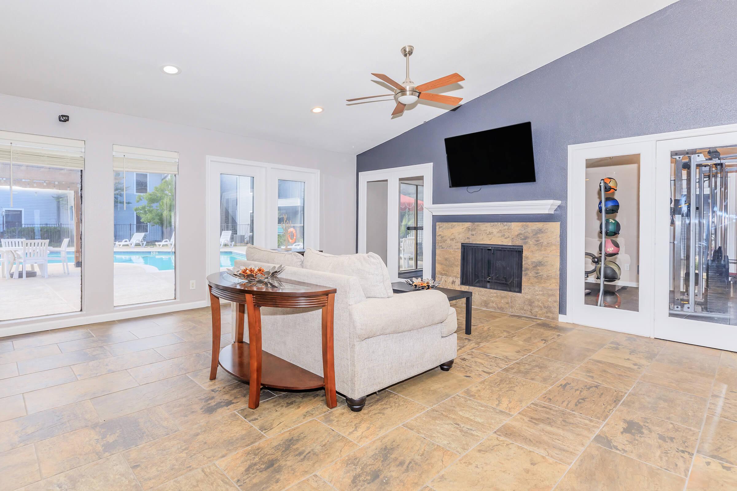 A modern living space featuring a light-colored couch and a round table, with large windows overlooking a pool area. The room includes a ceiling fan, a mounted TV, and a decorative fireplace, surrounded by neutral-toned tiled flooring. Fitness equipment is visible through glass doors.