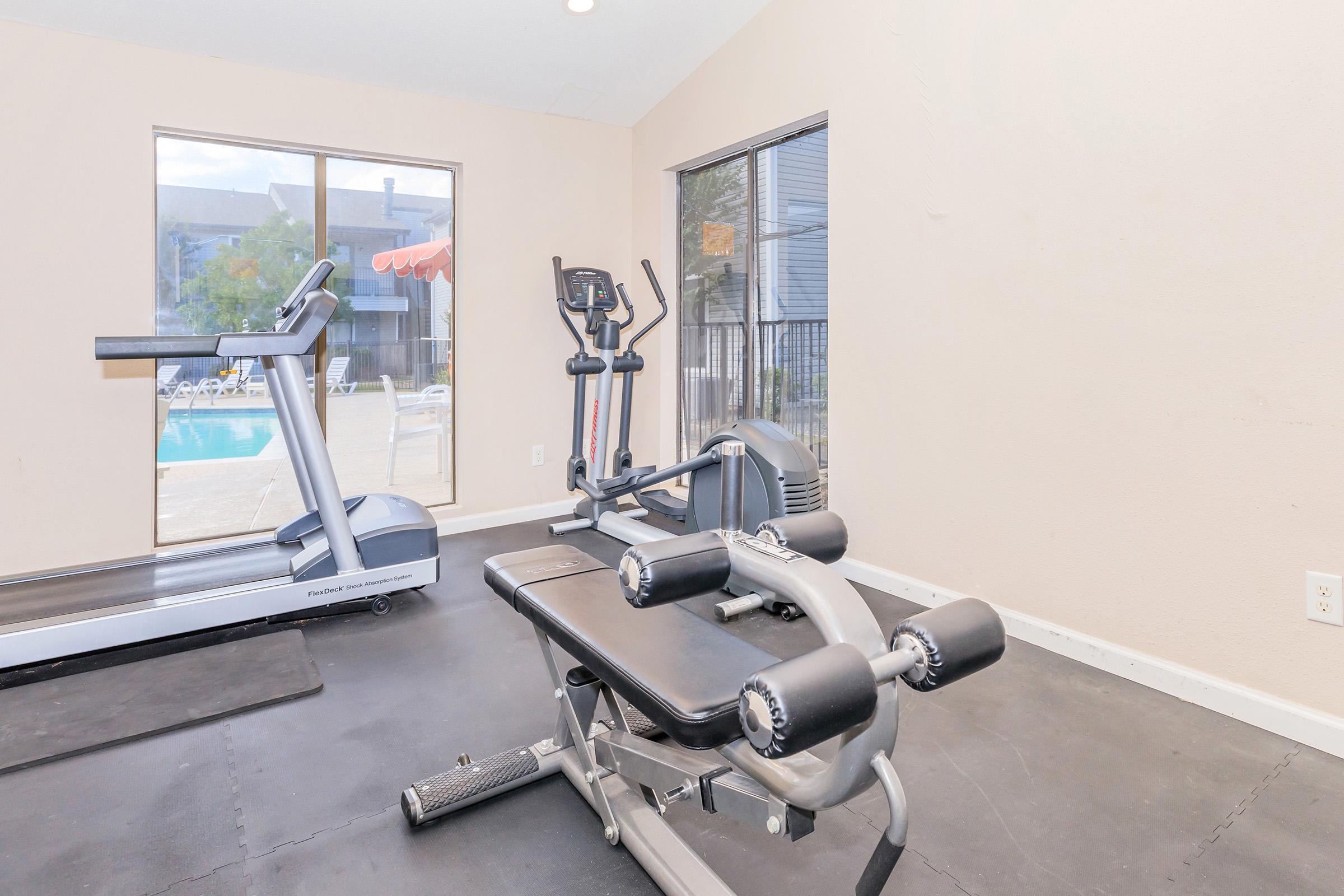 A well-lit gym with exercise equipment, including a treadmill, elliptical machine, and a workout bench with weights. In the background, large windows overlook a pool area, providing a view of outdoor space. The floor is dark and the walls are light-colored.
