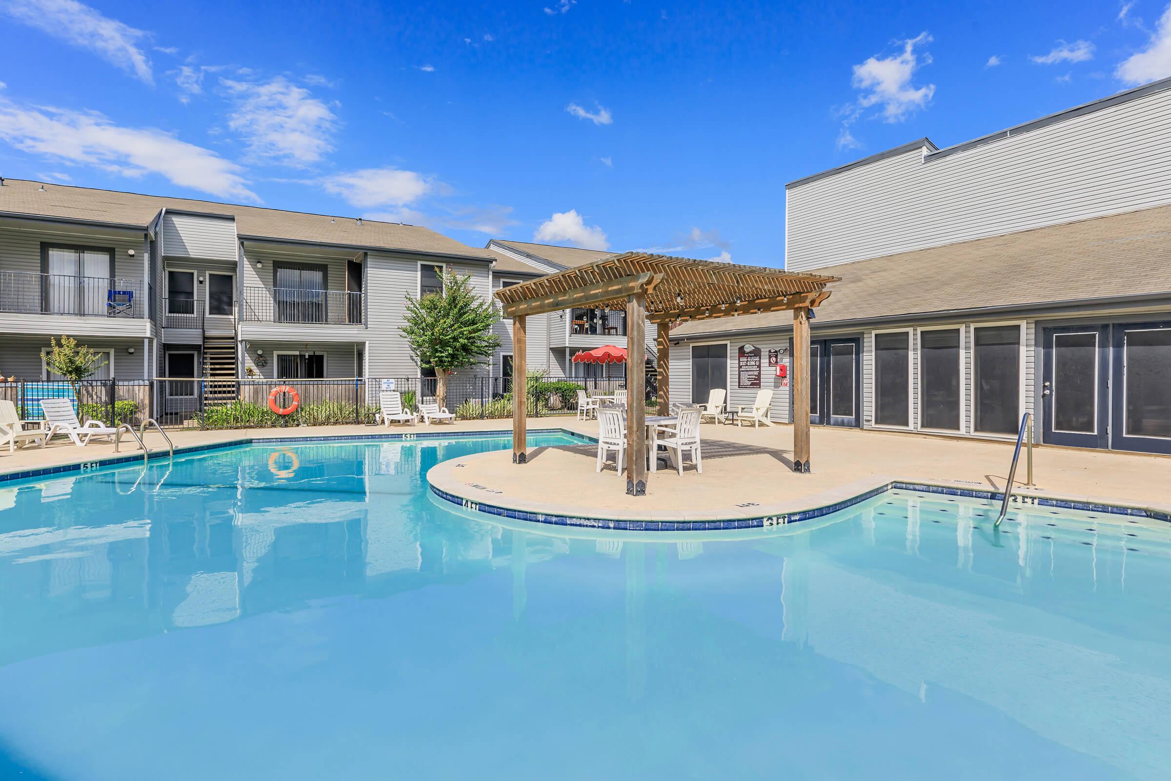 A sunny outdoor pool area with a wooden pergola and lounge chairs. The pool has clear blue water, and there are nearby apartment buildings surrounded by grass and landscaping. A few umbrellas provide shade, creating a relaxing atmosphere for residents.