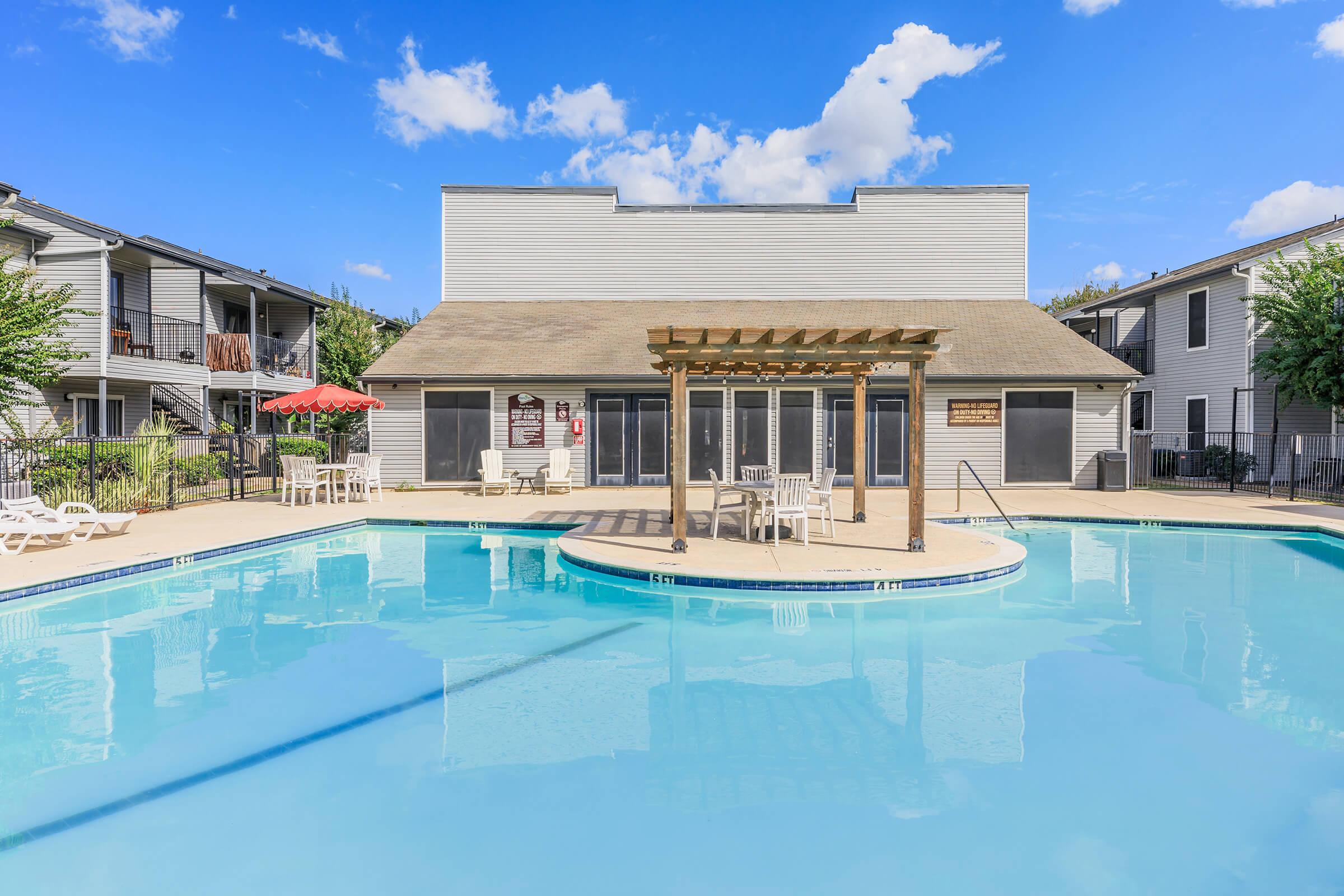 A clear blue swimming pool with a wooden pergola in the center, surrounded by lounge chairs and tables. In the background, a modern building with sliding glass doors and shaded seating areas. The sky is bright with fluffy white clouds.