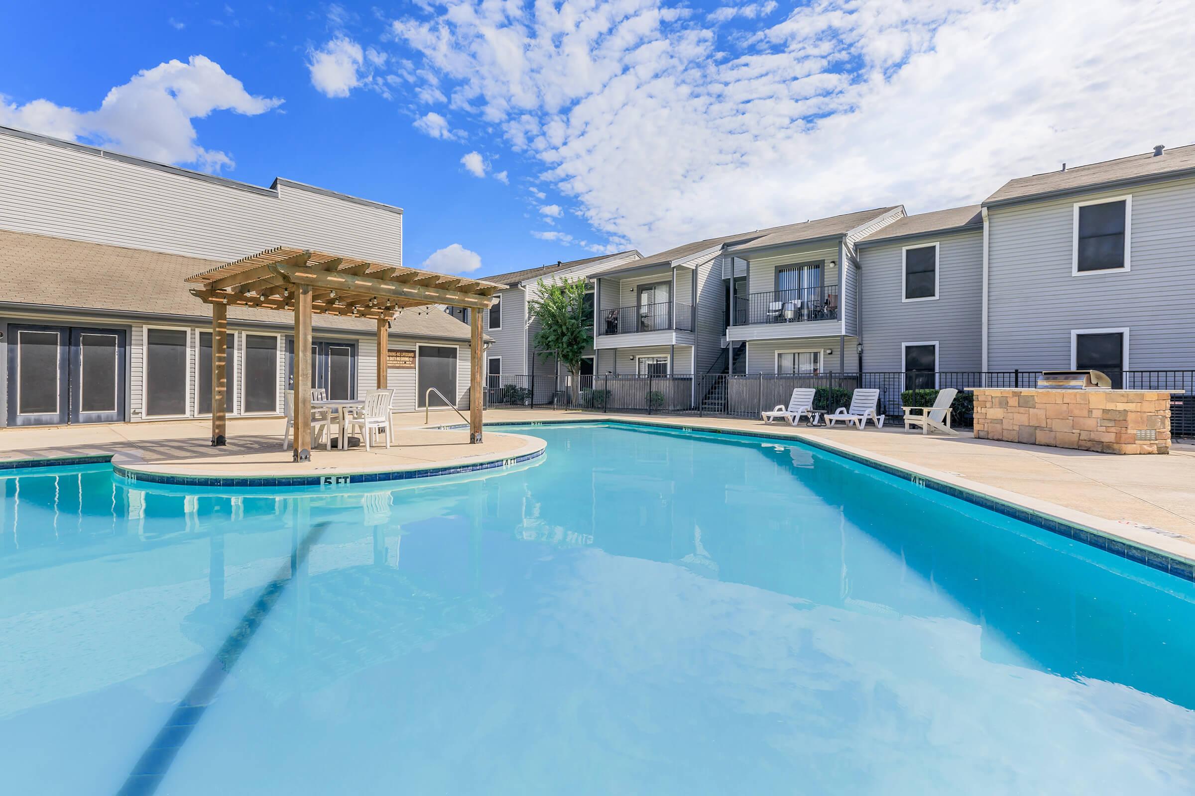 A clear blue swimming pool with a circular wooden pergola and seating area in the foreground. In the background, two multi-story apartment buildings are visible under a partially cloudy sky. Sunlight reflects off the water, creating a serene and inviting atmosphere.