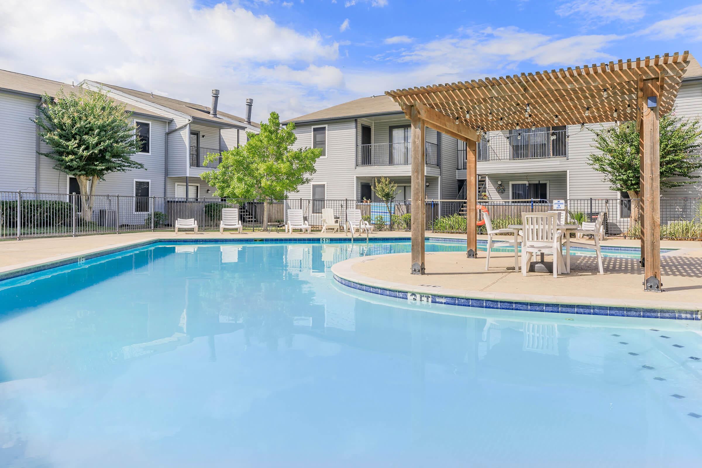 A clear blue swimming pool surrounded by a deck with lounge chairs. In the background, there are two-story apartment buildings and trees. A shaded pergola is situated near the pool, providing a relaxing spot for residents. The sky is bright with some clouds.