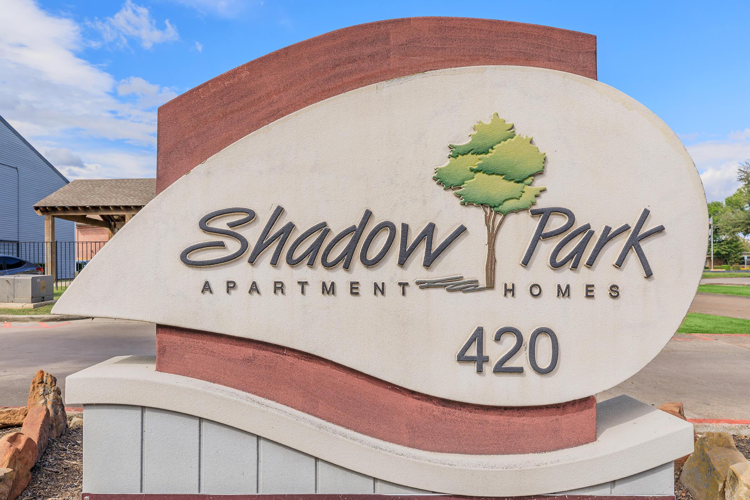 Sign for Shadow Park Apartment Homes with the number "420" displayed. The sign features a stylized design with a tree graphic and a blend of earthy colors against a blue sky backdrop.