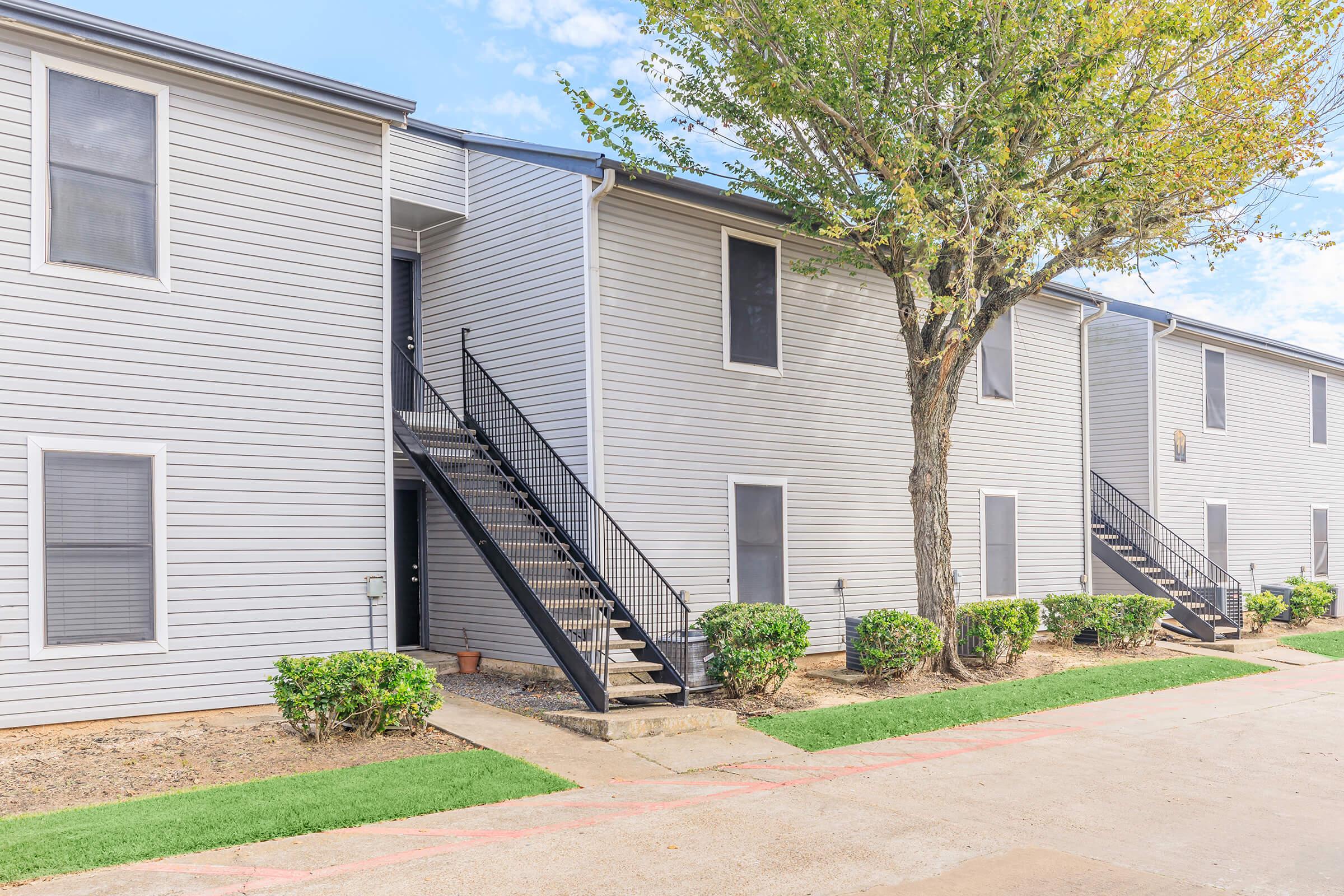 Two-story apartment building with gray siding and black metal staircases leading to the upper level. The landscaping includes small bushes and a tree, with a concrete pathway in front. The sky is clear with a few clouds.
