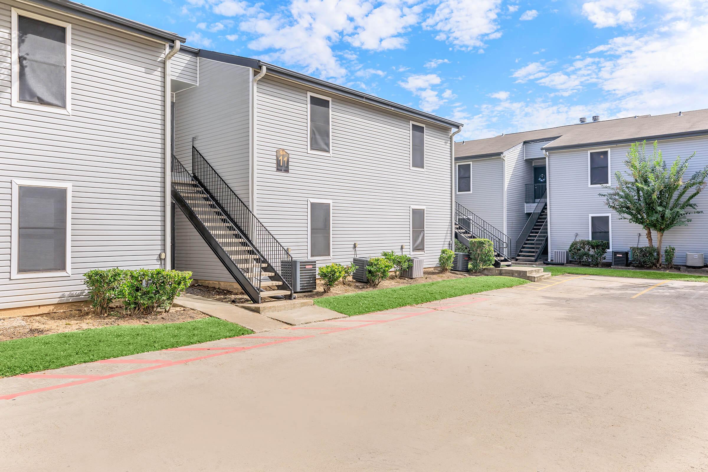 Two multi-story apartment buildings with gray siding and staircases leading to second-floor units. The foreground features a paved parking area with landscaped grass and small shrubs, while the background shows a clear blue sky with scattered clouds.
