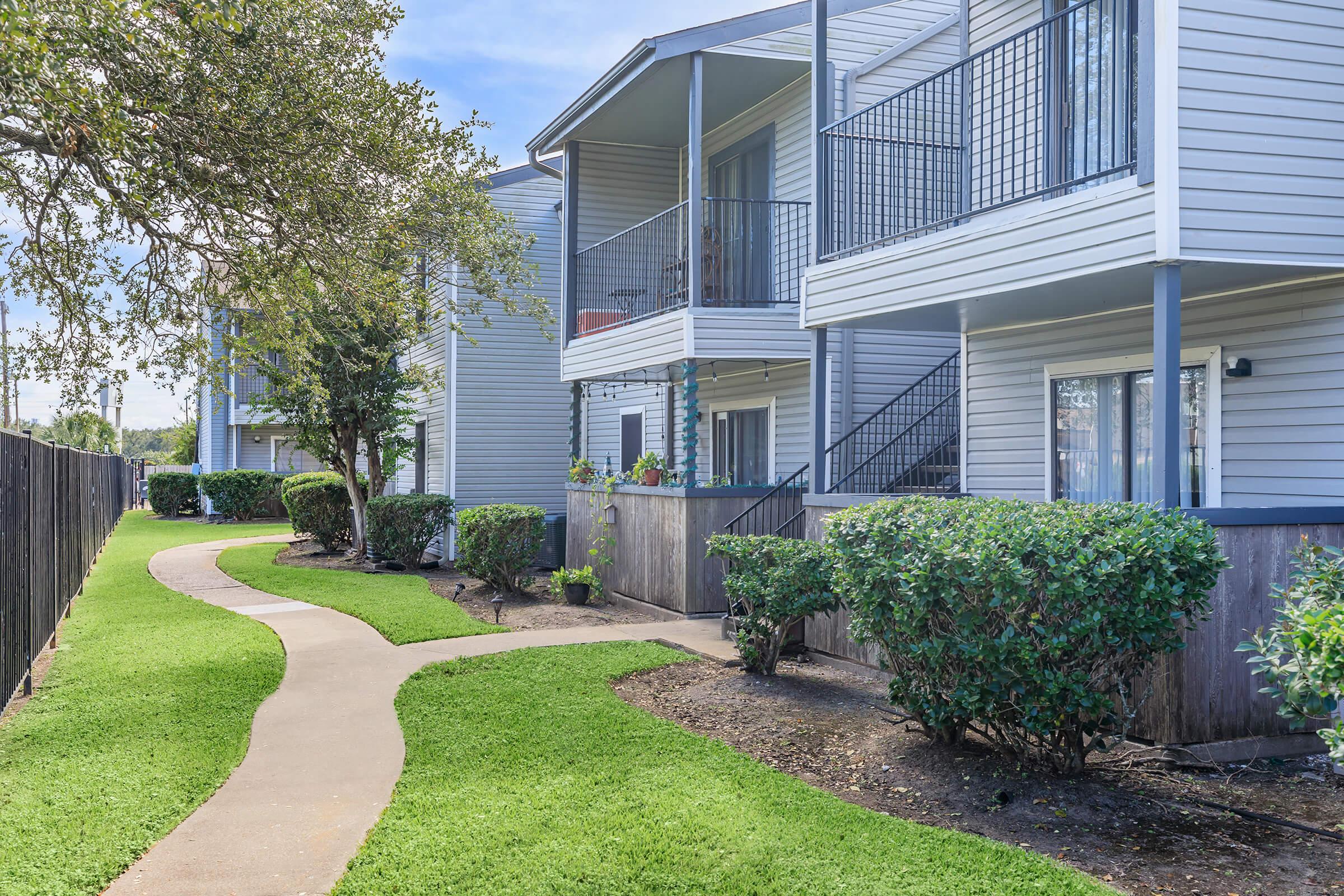 Pathway winding through a landscaped area with well-maintained grass and shrubs, leading to two-story apartment buildings with balconies. The buildings are painted in light colors and have metal railings, with a clear blue sky in the background.