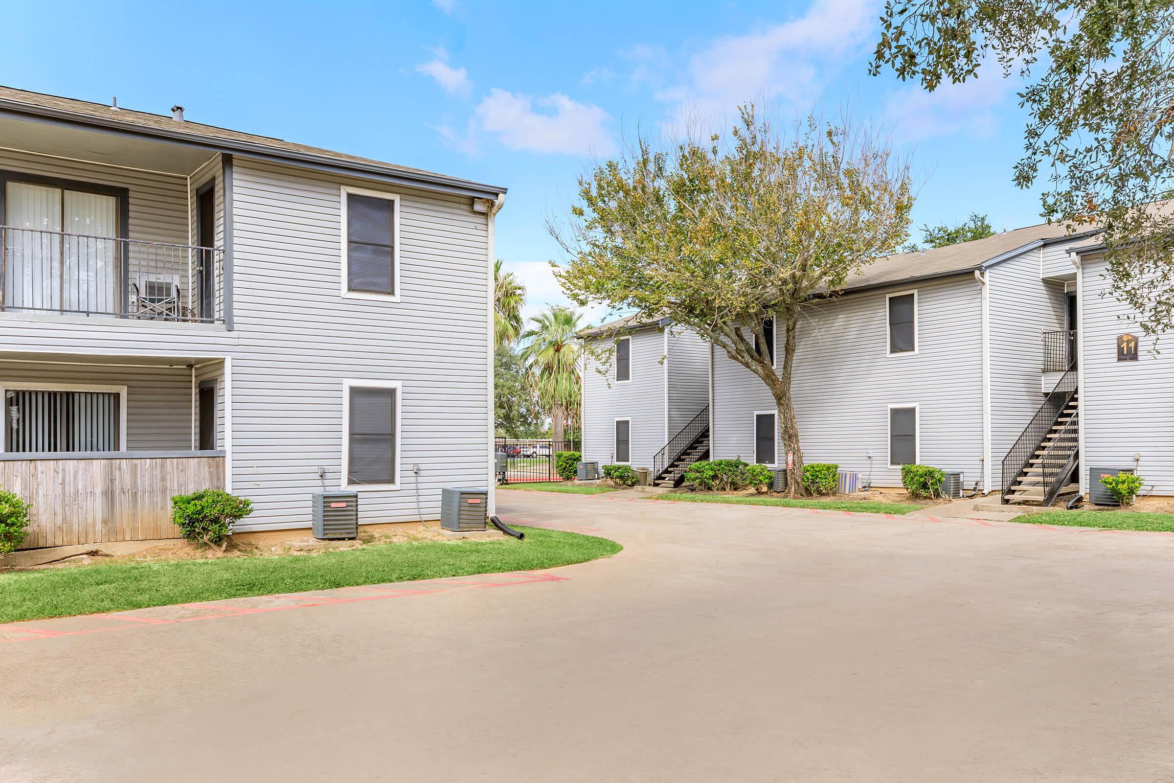Two multi-story apartment buildings with gray exteriors, surrounded by greenery. A tree is visible in the center, and there is a clear pathway leading through the area. The sky is partly cloudy, creating a bright atmosphere. Air conditioning units are placed near the ground level of the buildings.