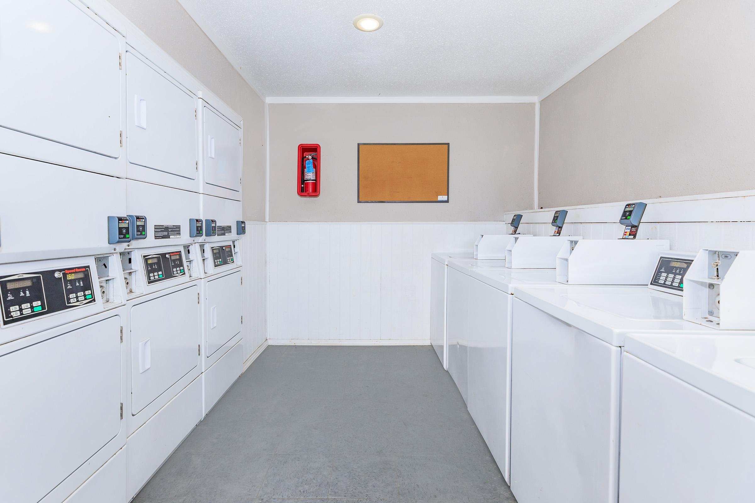 A clean and organized laundry room featuring several stacked white washing machines and dryers along the left wall, digital controls visible on each unit. A bulletin board is mounted on the wall, and a fire extinguisher is visible for safety. The floor is grey and the walls are light-colored.