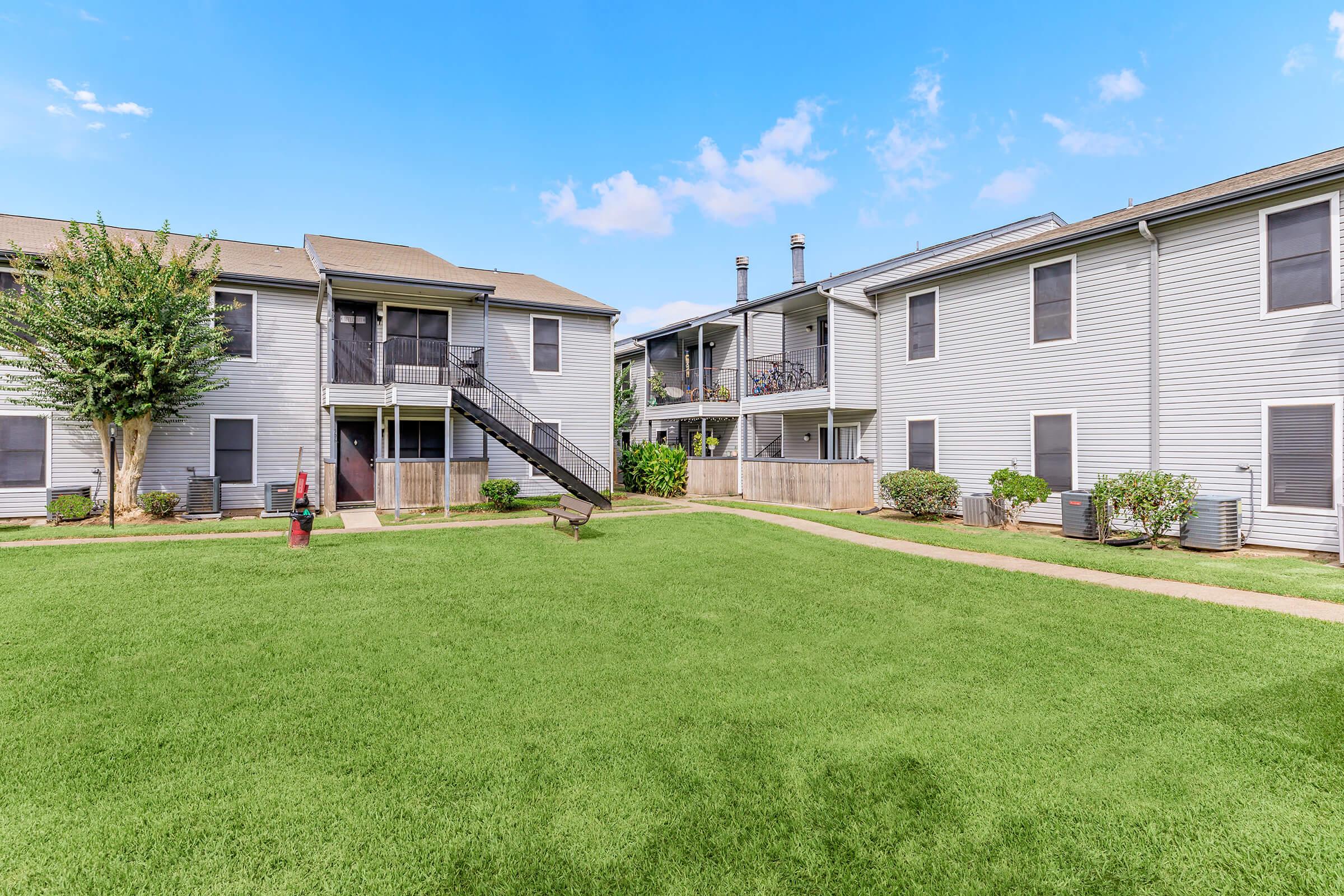 A view of a well-maintained apartment complex featuring two buildings. One building has stairs leading to upper units, while the other has a balcony. The green lawn is manicured, with small shrubs and trees adding greenery, and pathways connecting the areas, under a clear blue sky.
