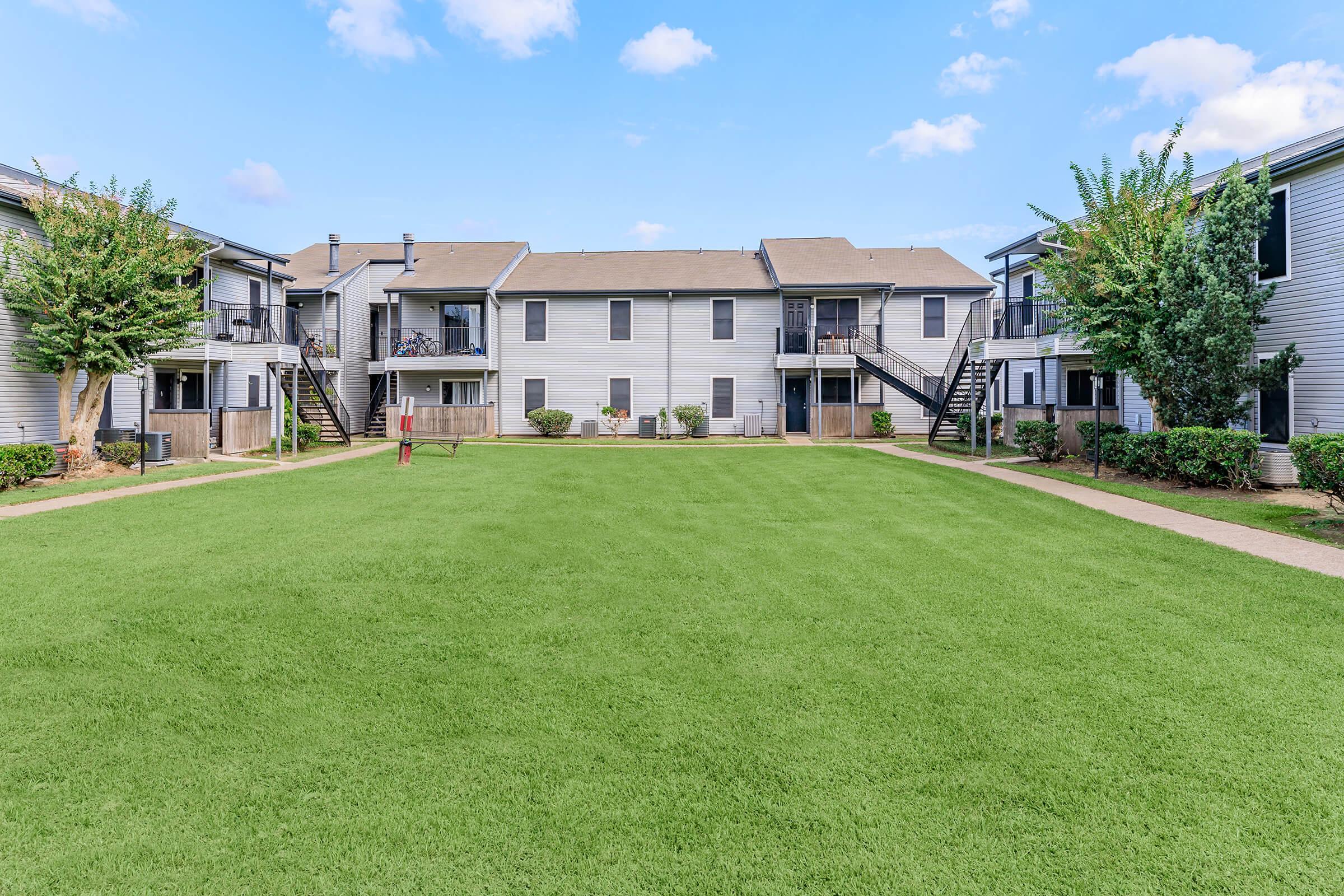 A well-maintained apartment complex featuring two-story buildings with balconies. In the foreground, a lush green lawn stretches between the buildings, surrounded by landscaping and trees. The sky is clear with a few clouds visible.