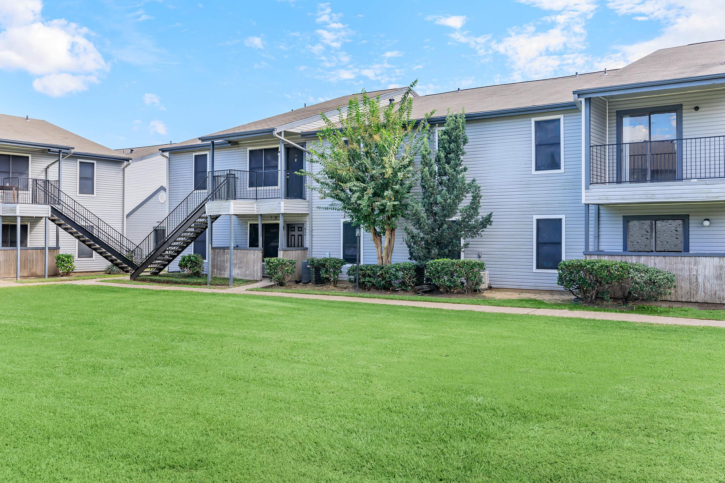A modern apartment complex featuring two-story buildings with balconies, surrounded by well-maintained lawns and shrubbery. A set of stairs is visible leading to the second floor of an apartment. The sky is clear with a few scattered clouds.