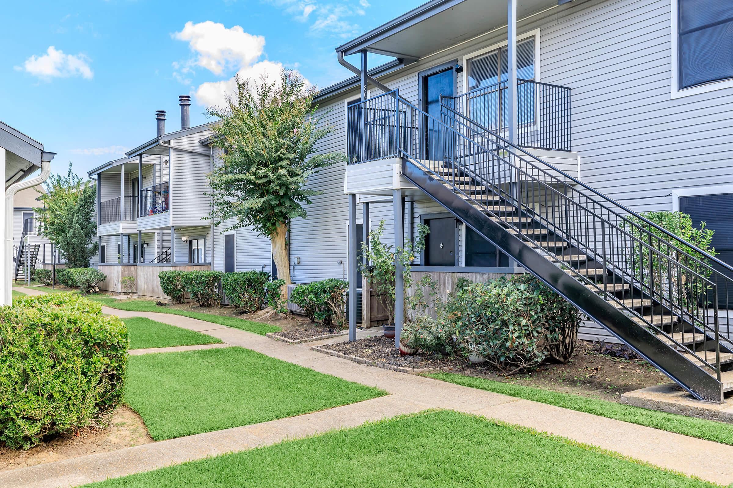 A view of a residential apartment complex featuring two-story buildings with a staircase leading to an upper-level unit. Lush green lawns and neatly trimmed hedges line the pathways, creating a well-maintained appearance. The sky is clear with a few clouds, enhancing the pleasant outdoor setting.