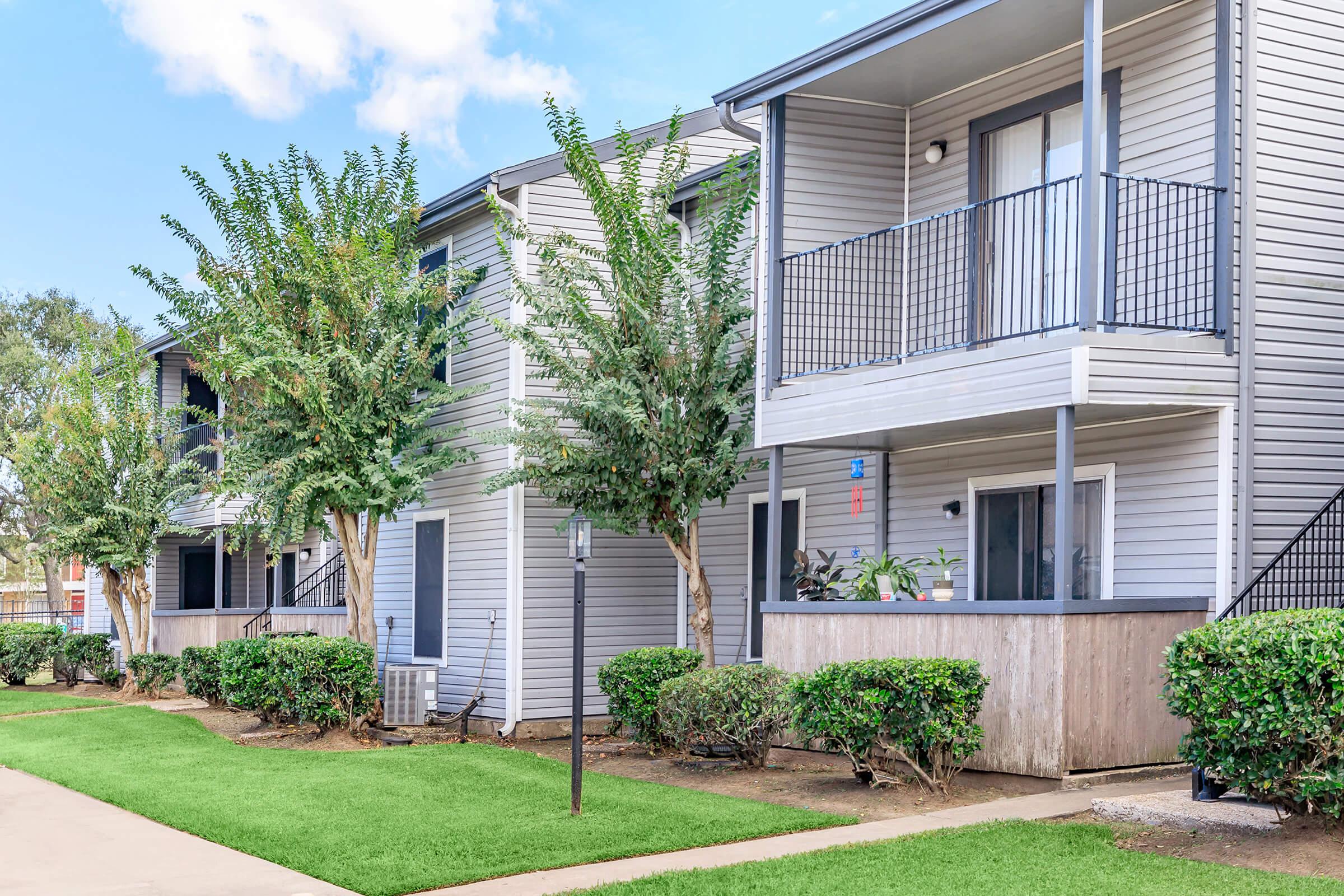 A row of modern apartment buildings with gray siding, featuring balconies and surrounded by well-maintained greenery. The lawn is neatly trimmed, and trees provide shade. The scene is bright and inviting, with a clear blue sky in the background.