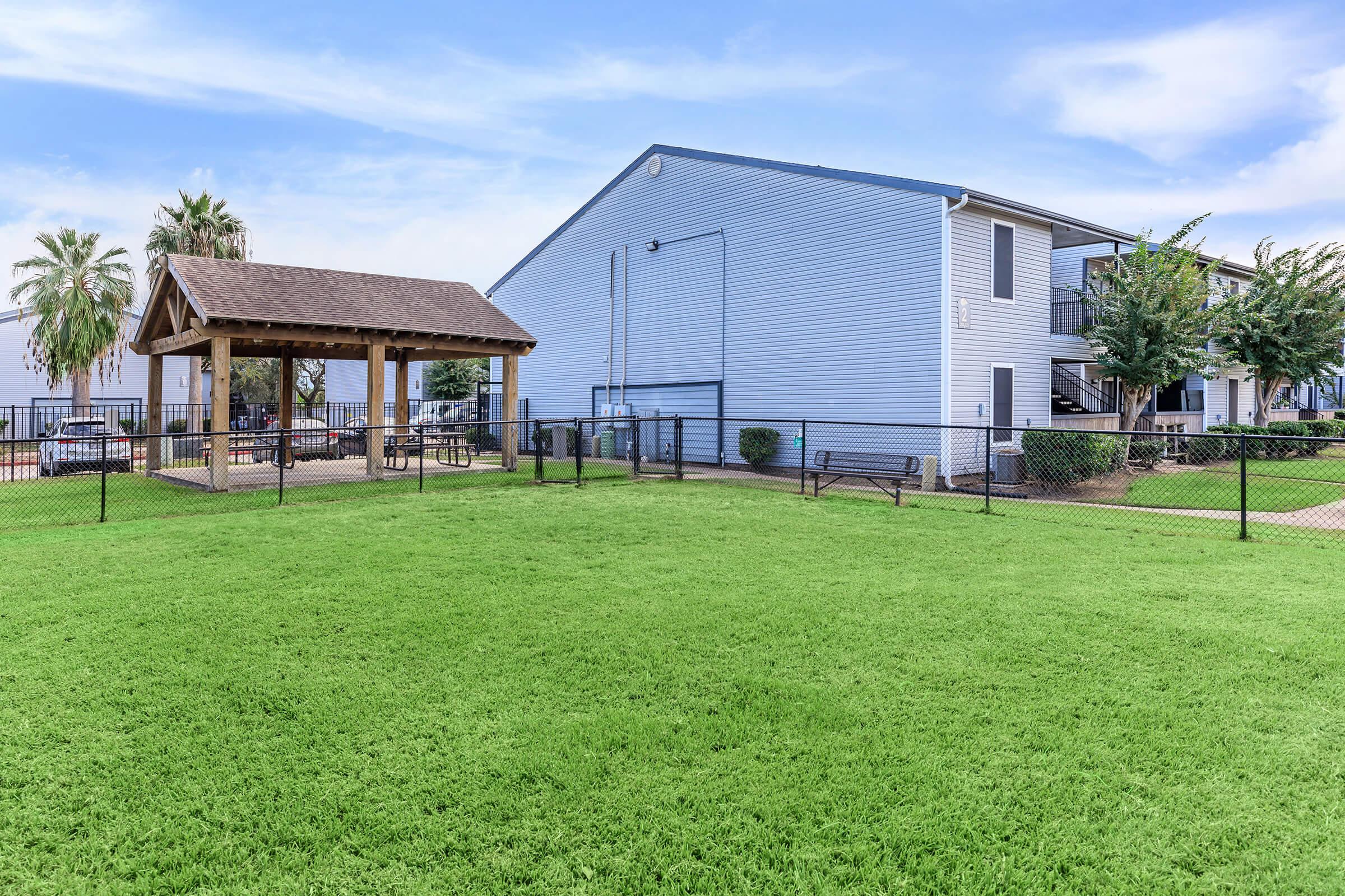 A well-maintained green lawn in front of a residential building. A wooden gazebo with picnic tables is visible, along with a fenced area, palm trees, and the exterior of a blue apartment complex. The sky is clear with a few clouds, creating a bright and inviting atmosphere.