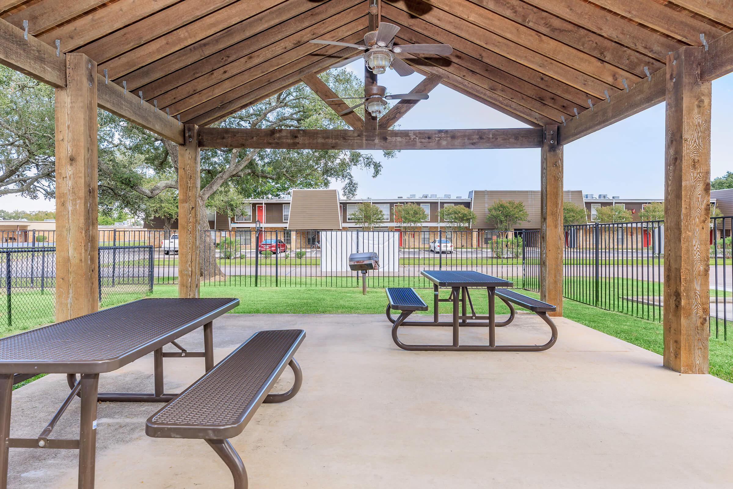 A shaded outdoor pavilion with a wooden frame and a ceiling fan, featuring picnic tables and a grill. The pavilion overlooks a grassy area and a parking lot, with several buildings visible in the background. The setting is well-maintained and inviting for gatherings or relaxation.