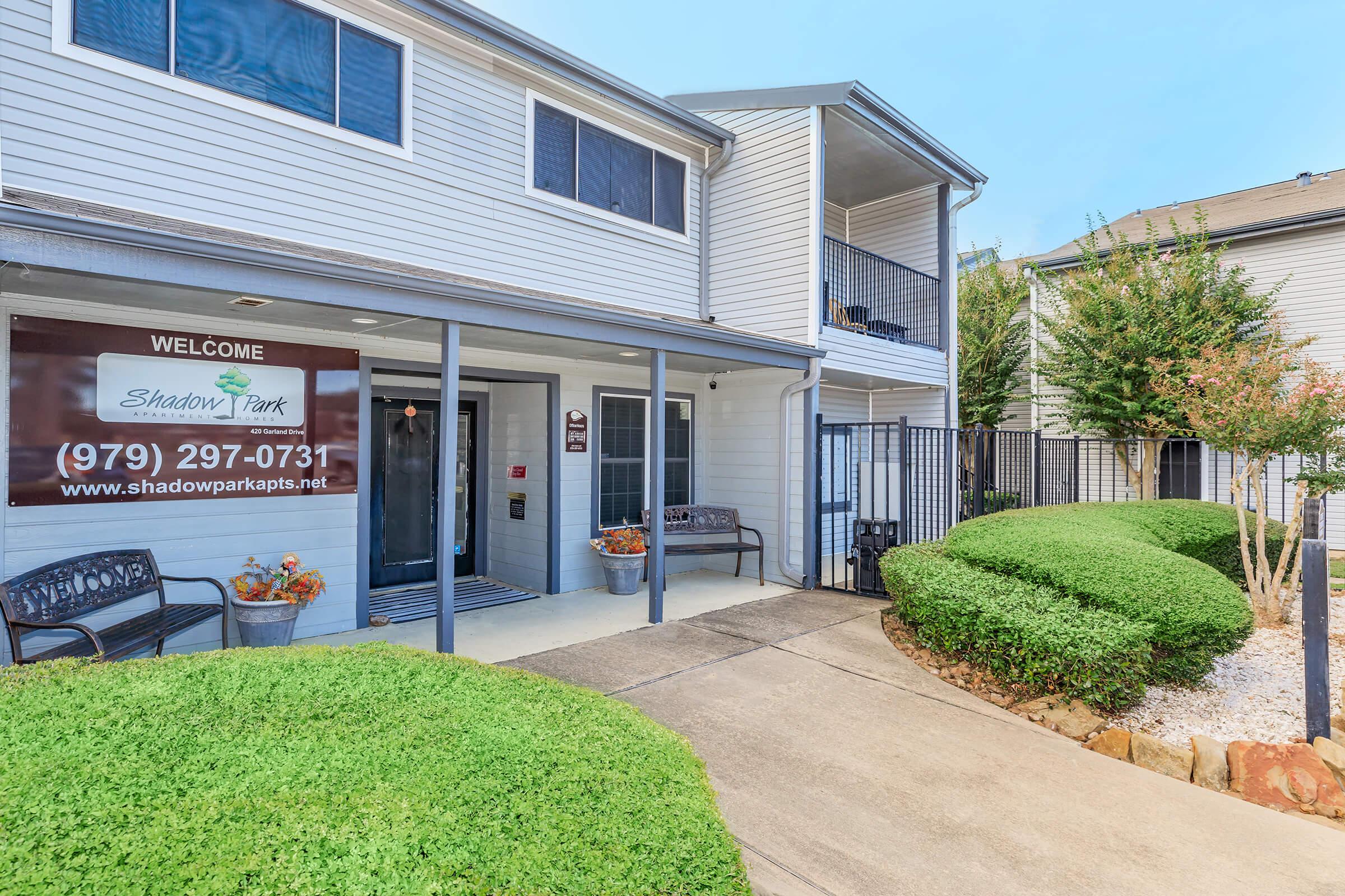 Exterior view of an apartment complex entrance featuring welcoming signage, well-maintained landscaping with trimmed hedges, and a pathway leading to the entrance. The building has two stories with balconies, and a decorative seating area is visible in front.