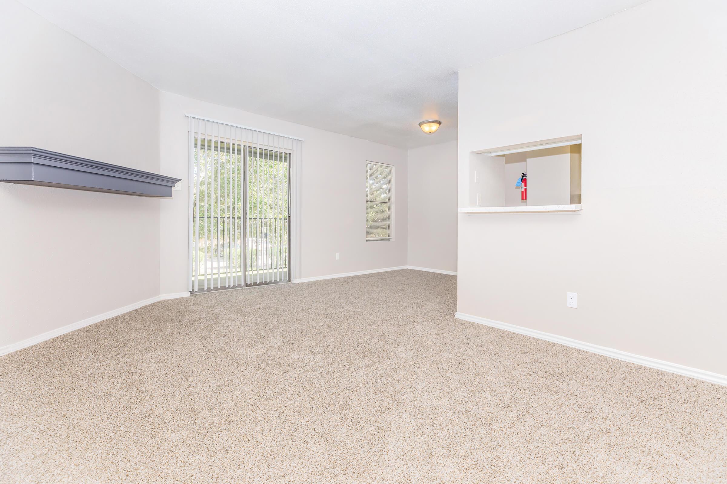 A spacious, empty living room featuring light beige carpet and neutral-colored walls. Large windows with vertical blinds allow natural light. A small serving bar is visible in the background, separating the living area from the adjacent kitchen.