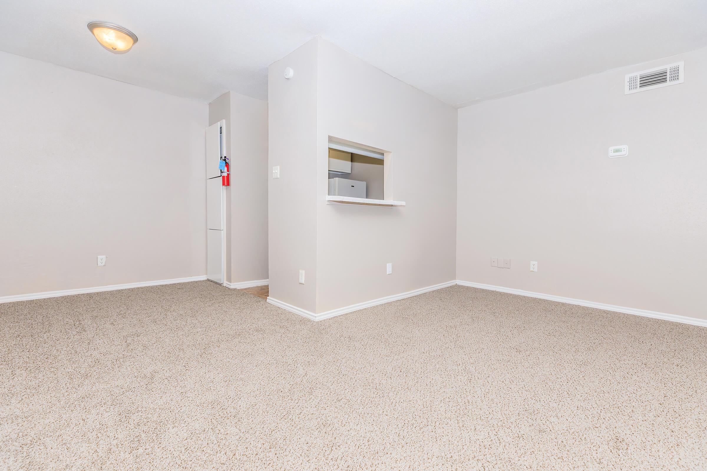 A spacious, empty living area with light beige walls and carpet. There is a small opening that leads to a kitchen, visible in the background with a white refrigerator. The ceiling has a light fixture, and a wall-mounted thermostat is present. The overall atmosphere is bright and clean.