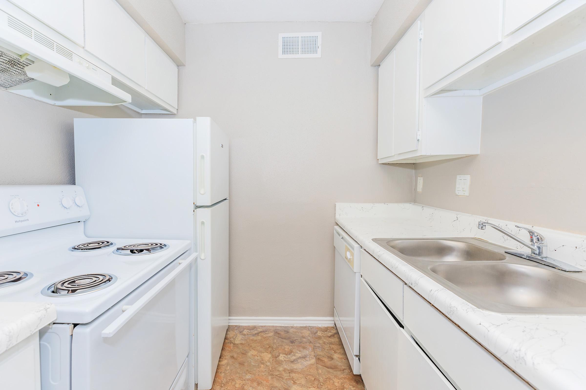 A small, compact kitchen featuring white appliances including a stove, refrigerator, and double sink. The countertops are light-colored with a marbled design, and the walls are painted in a neutral tone. The space appears tidy and well-organized, with cabinets above the counters for storage.