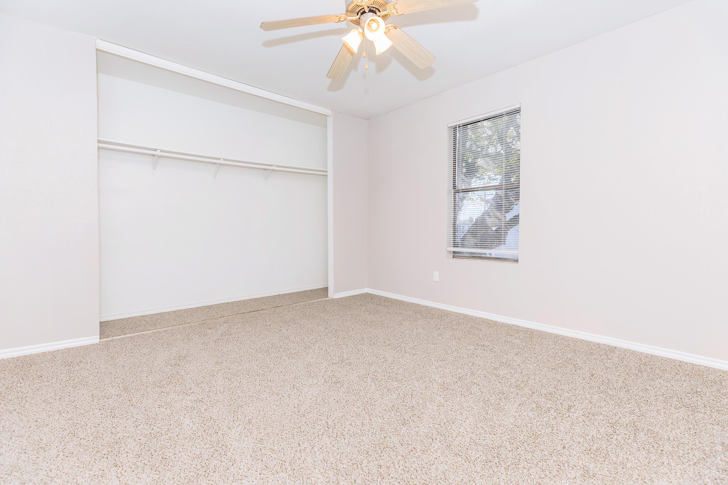 A spacious, empty room with light beige carpet and a ceiling fan. There is a large window with blinds allowing natural light, and a built-in closet with a shelf. The walls are painted a light color, contributing to a bright and airy atmosphere.