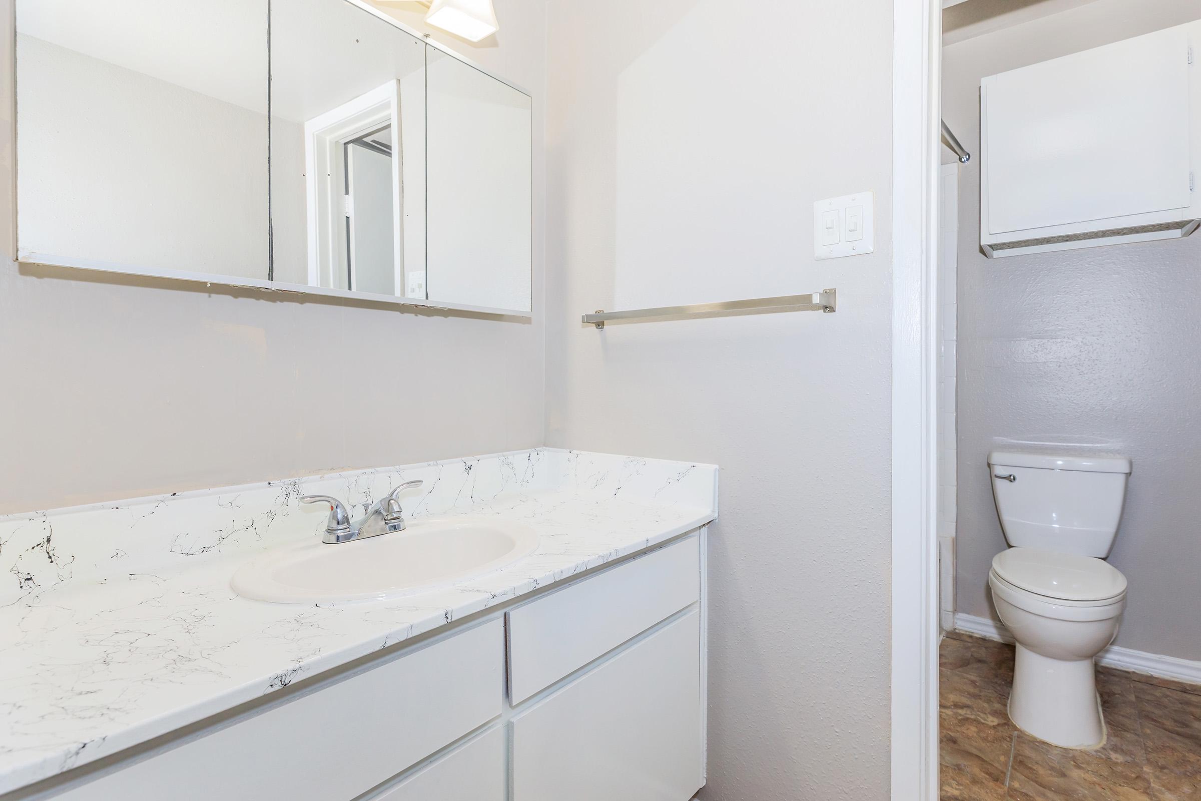 A clean, modern bathroom featuring a white countertop with a sink, a large mirror above, and light-colored walls. There is a towel rack and a toilet in the corner, with textured flooring completing the minimalist design.
