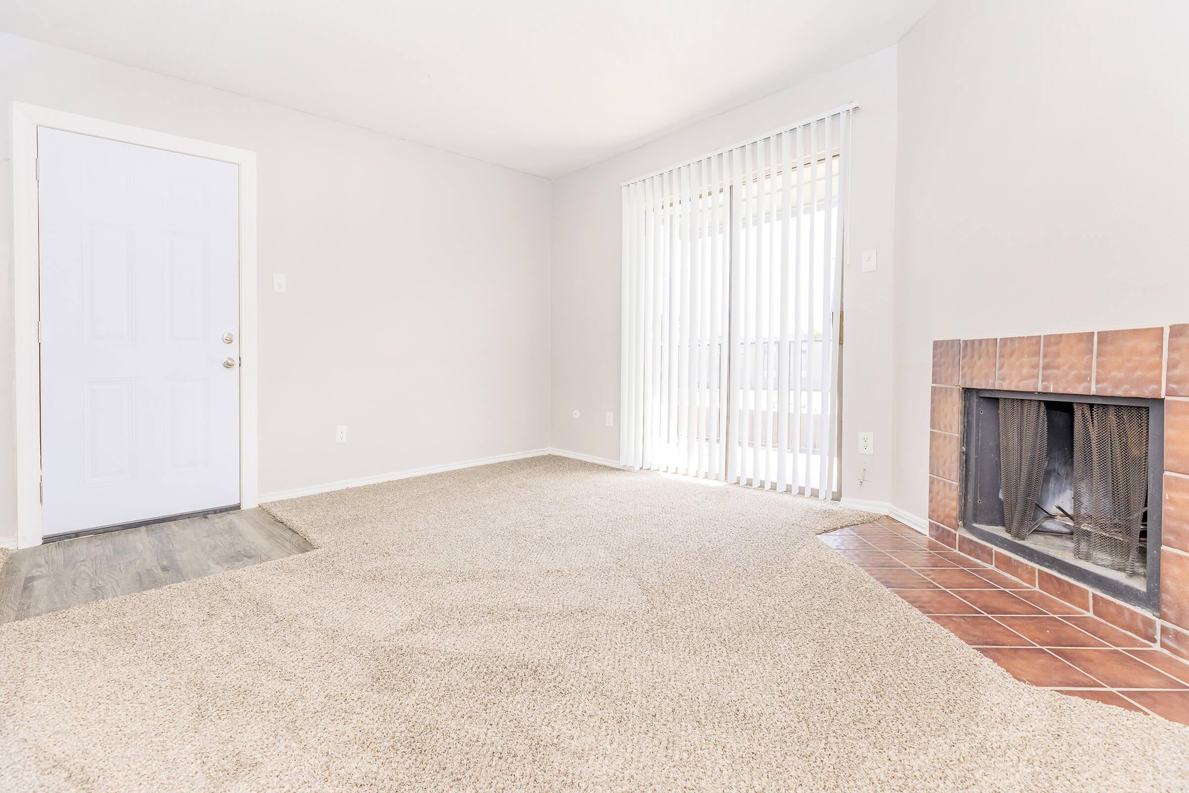 A spacious and empty living room featuring light gray walls, beige carpet, a door leading outside, and a sliding glass door with vertical blinds. There's a tiled fireplace on one wall, creating a cozy atmosphere. The room is well-lit, emphasizing its openness and potential for decoration.