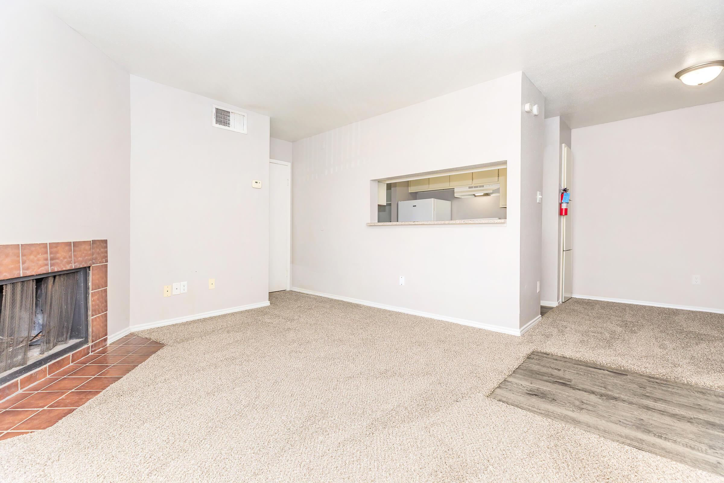A spacious, empty living room with light gray walls and carpet. A fireplace is on the left with a tiled surround. In the background, there's a partial view of the kitchen through an opening in the wall. The room is well-lit and free of furniture, creating a clean and open atmosphere.