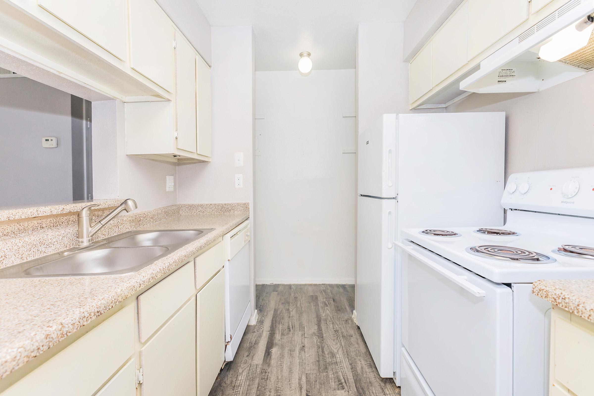 A clean, modern kitchen featuring light-colored cabinets, a double sink, a white refrigerator, and a white stove. The countertops are a speckled beige, and the flooring is a light wood laminate. The space is well-lit with a ceiling light fixture, and there's a neutral-colored wall in the background.