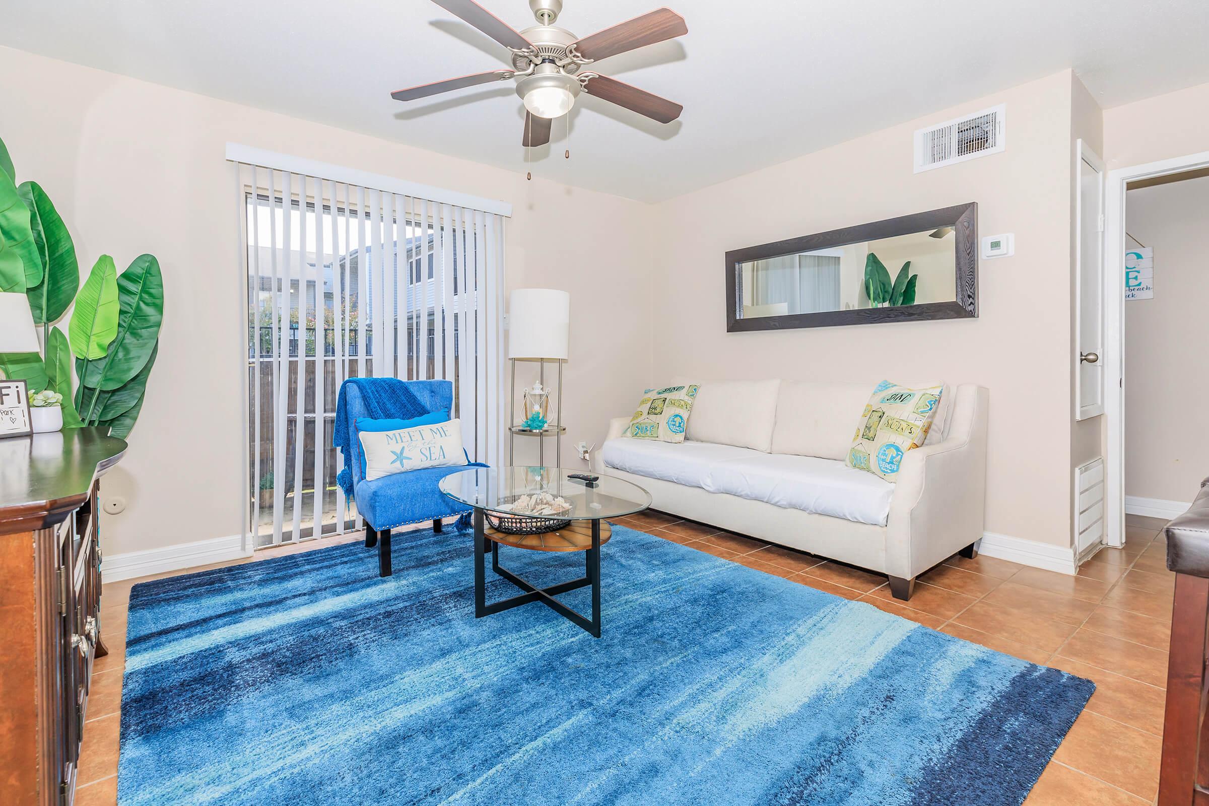 A cozy living room featuring a blue area rug, a white couch with decorative pillows, a blue accent chair, a glass coffee table, and a ceiling fan. Large windows with retractable blinds allow natural light, and decorative plants add a touch of greenery. A mirror hangs on the wall, enhancing the space's brightness.