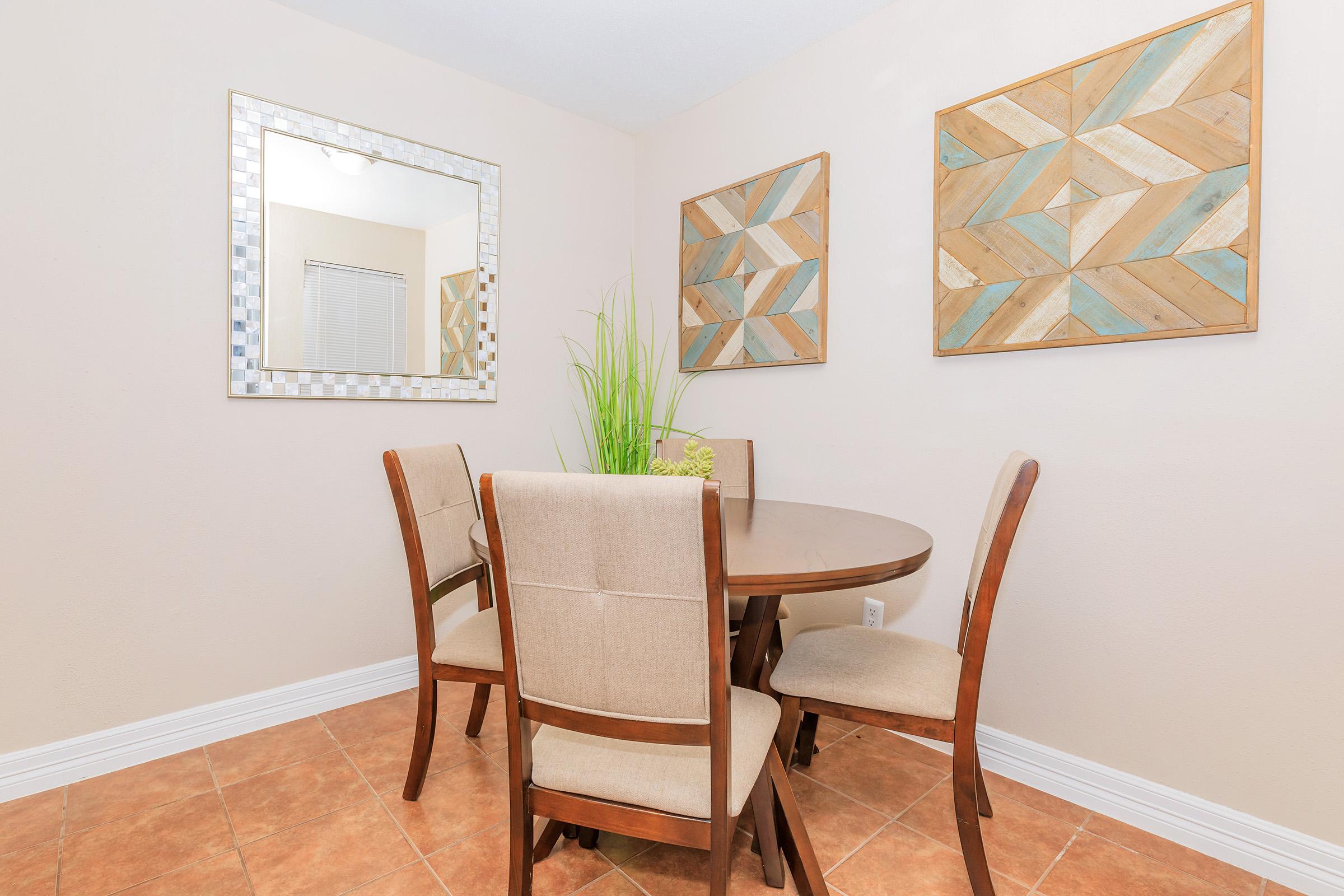 A cozy dining area featuring a round wooden table surrounded by four beige upholstered chairs. The walls are adorned with two geometric-patterned wooden wall art pieces, and a large mirror reflects the space. A decorative plant adds a touch of greenery to the setting.