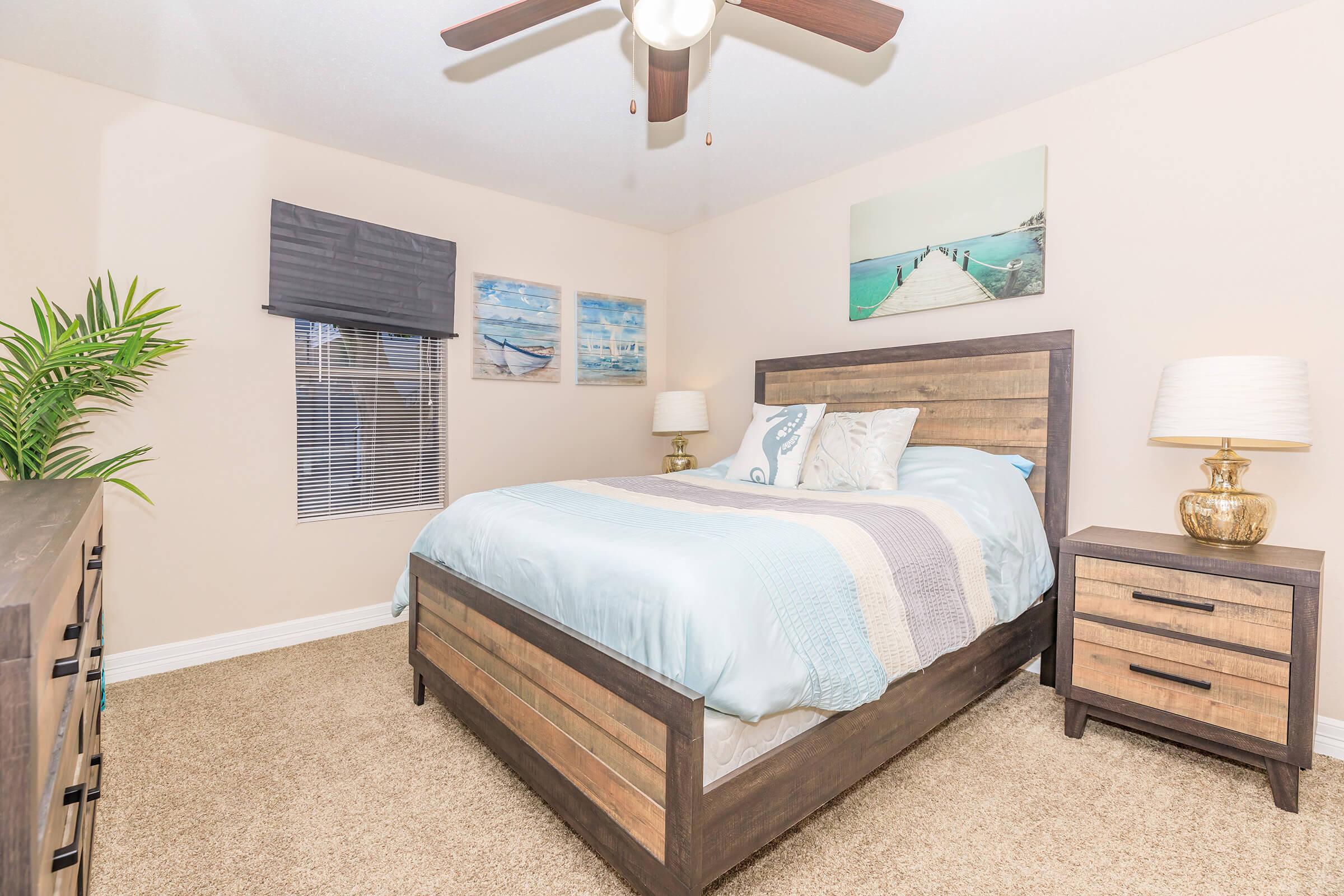 A cozy bedroom featuring a wooden bed frame with light blue and beige bedding. Adjacent nightstands with lamps flank the bed. A dresser is visible on the left, and a green plant adds a touch of nature. The walls are adorned with nautical-themed artwork, and the room has a ceiling fan and window coverings.