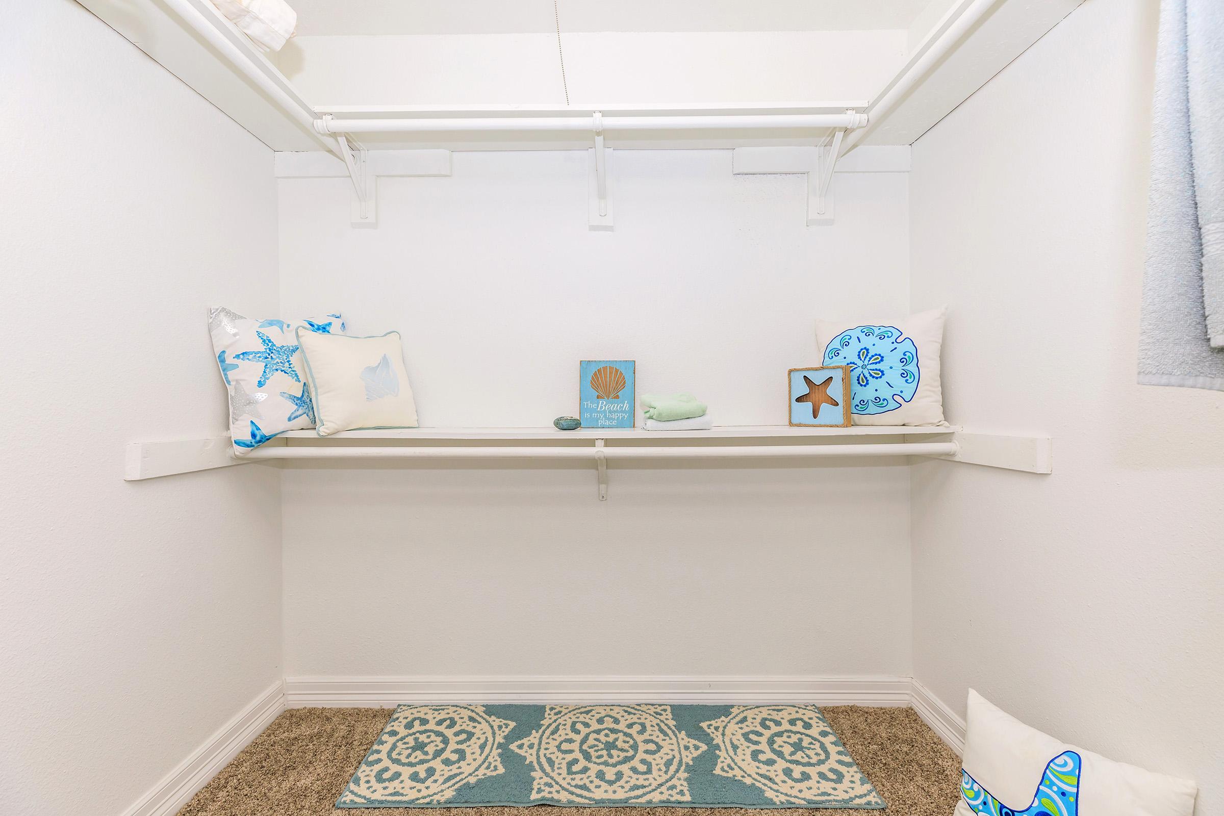 An empty closet with white walls and a light-colored carpet. It features two shelves with decorative pillows and a small framed picture, along with a light blue towel. The floor is adorned with a patterned rug, enhancing the inviting atmosphere of the space.