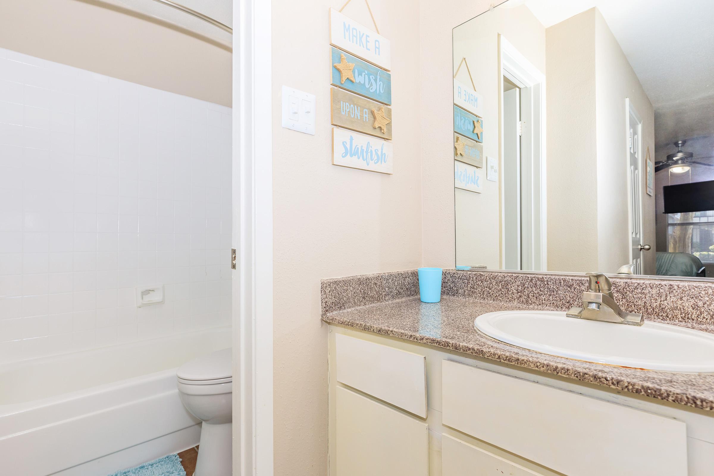Bright bathroom with a light-colored wall and a countertop featuring a sink. On the wall, there are two decorative signs. A white bathtub is visible in the background, and a light blue cup is placed on the countertop. The space is designed for functionality and comfort.