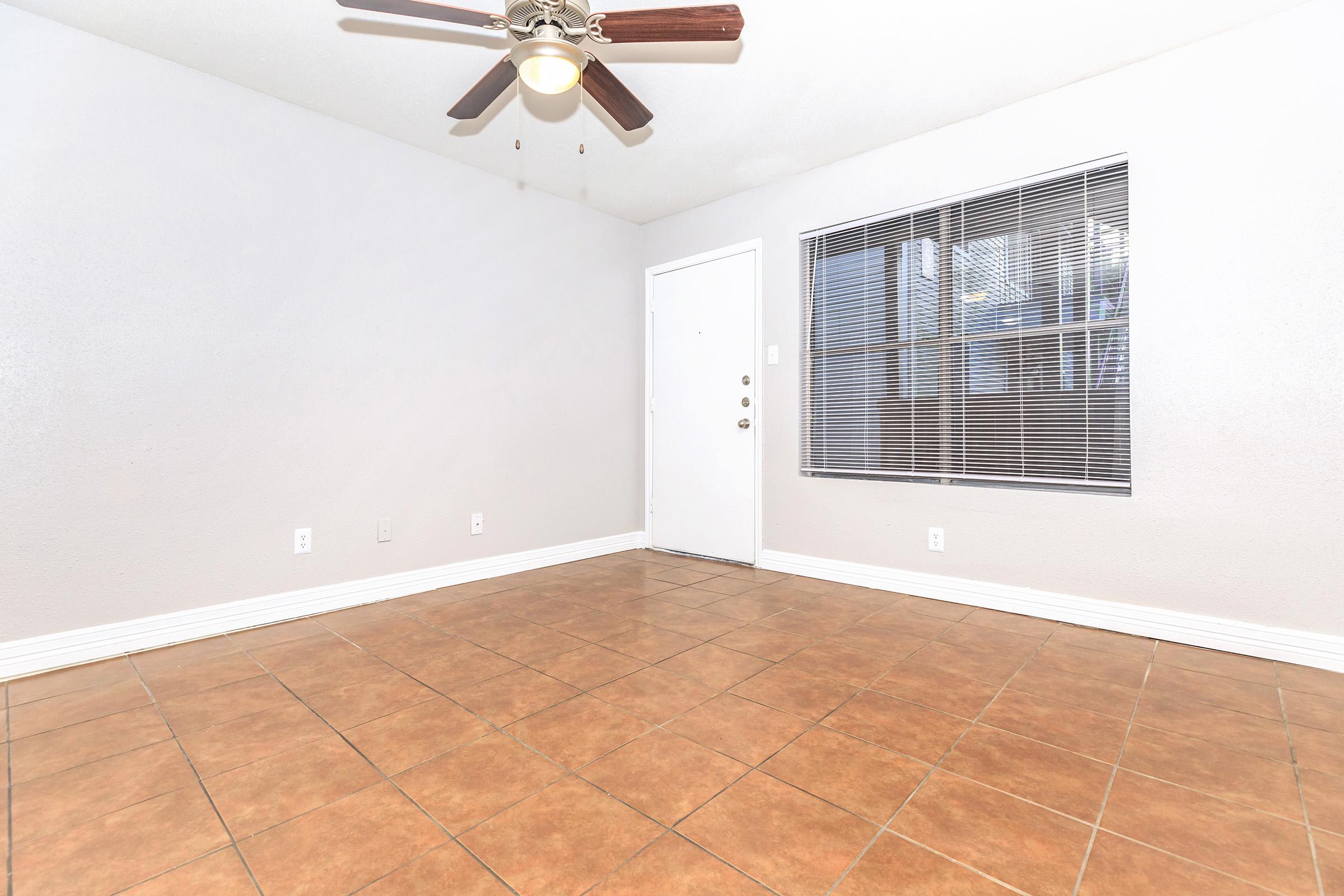 A spacious, empty room with brown tiled flooring, a ceiling fan with light, and a large window featuring blinds. The room has light gray walls and a white door, creating a bright and airy atmosphere. Ideal for future decoration or use as a living space.