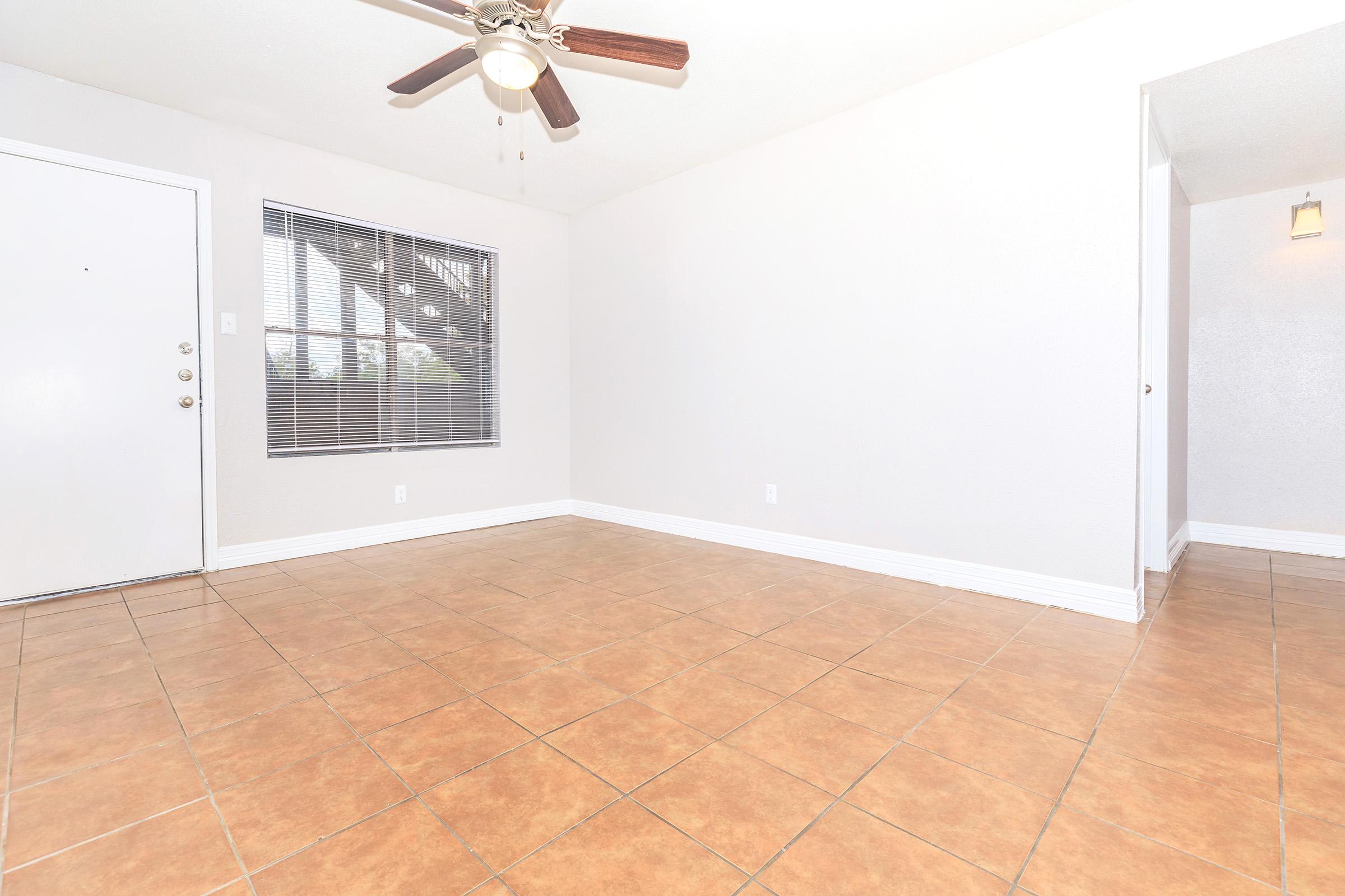 Spacious interior of an empty room with beige tiled flooring, a ceiling fan, and a window with blinds. The walls are painted light gray, and there’s an entrance door visible on the left side. The room is well-lit, giving a clean and inviting ambiance.