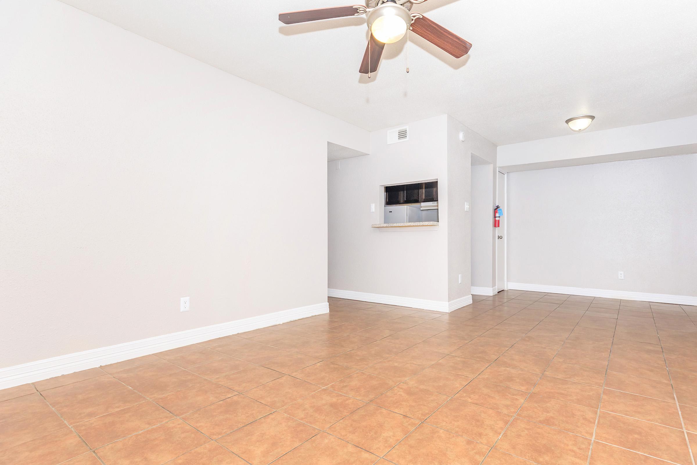 Spacious, empty living area featuring tiled flooring, a ceiling fan, and a neutral-colored wall. A small opening leads to another room, and there's a partial wall with an inset shelf. The area is well-lit, emphasizing the clean and open feel of the space.