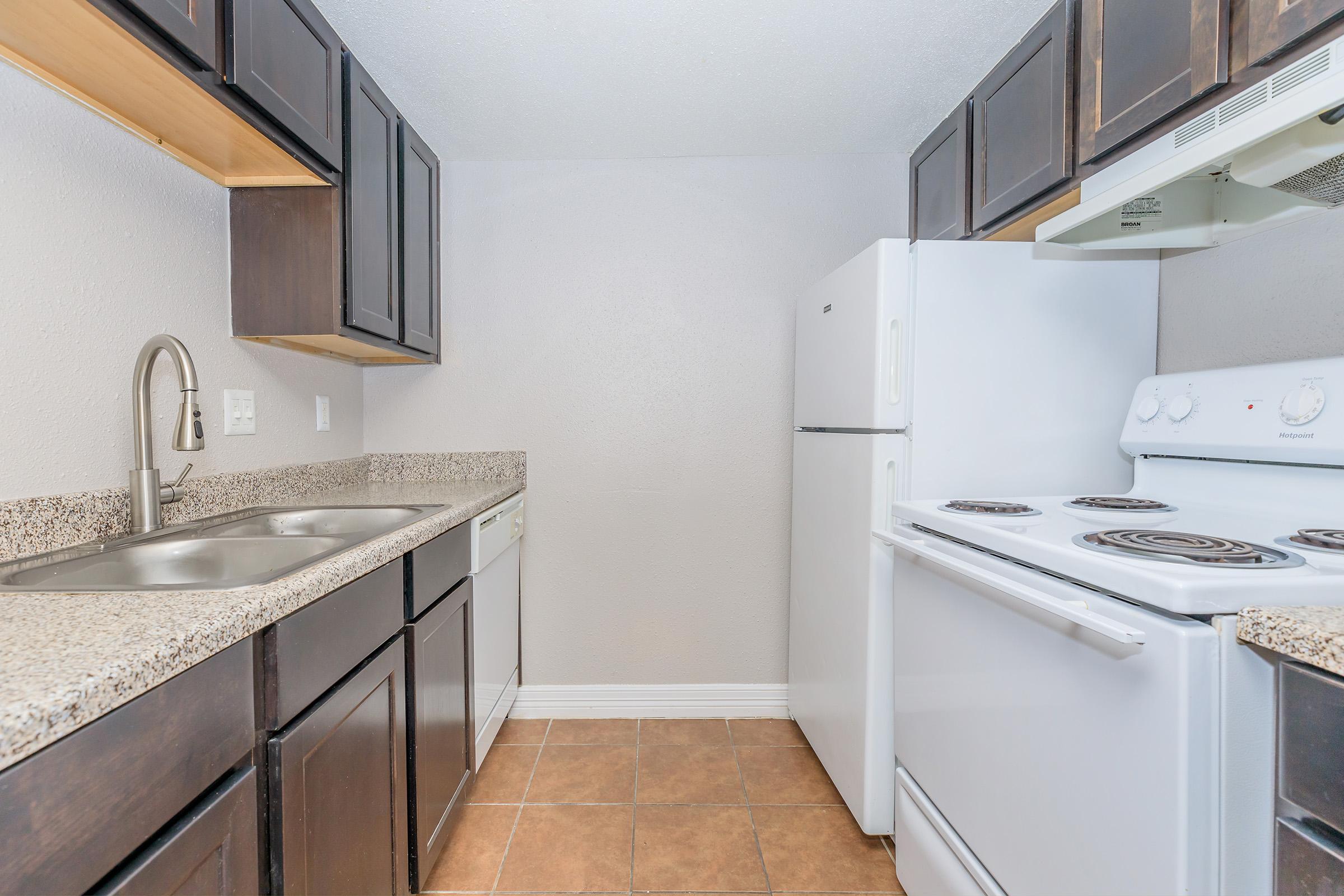A modern kitchen featuring dark wood cabinets, a granite countertop, a stainless steel sink, and white appliances including a refrigerator and stove. The walls are painted light gray, and the floor is tiled in a warm brown color, creating a clean and inviting atmosphere.