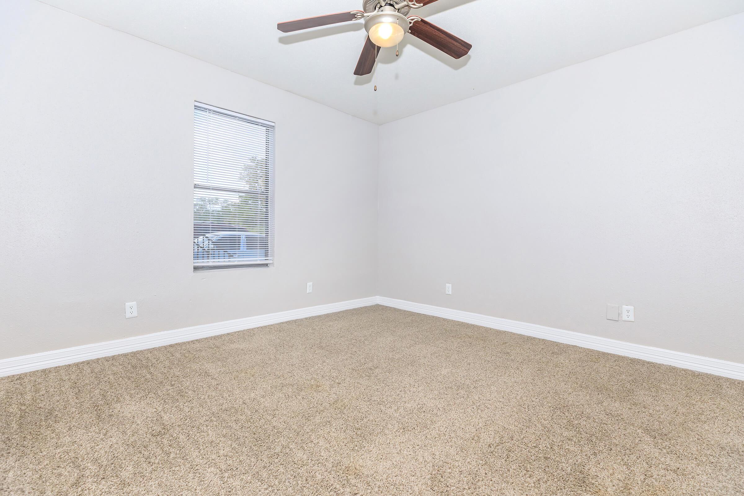 Empty room with light beige carpet, a ceiling fan with three blades, and a single window covered with blinds, allowing natural light to enter. The walls are painted in a light gray color, and there are no furnishings or decorations visible in the space.